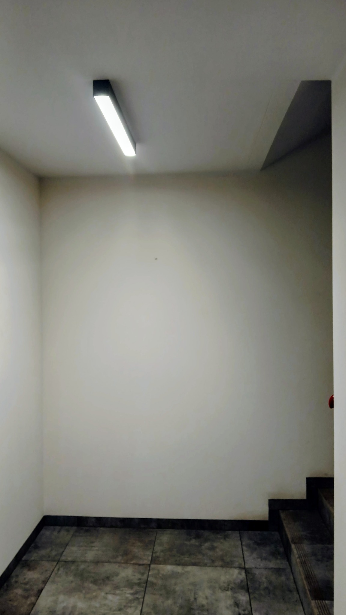 Pusta, jasna ściana w korytarzu z szarą podłogą z płytek i nowoczesnym oświetleniem sufitowym. Widoczne schody w tle.