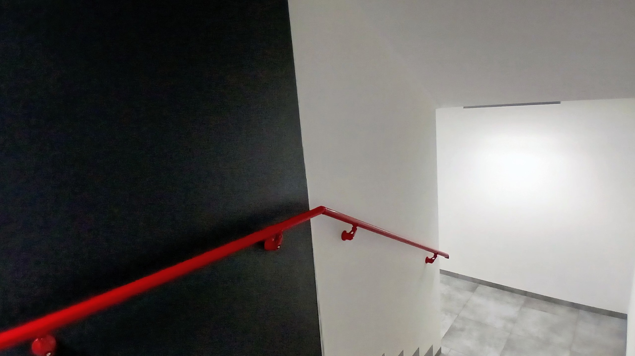 Klatka schodowa po remoncie: czarna ściana kontrastuje z białą, a czerwona poręcz dodaje akcentu kolorystycznego. Widoczne płytki na podłodze i świeżo pomalowane ściany.