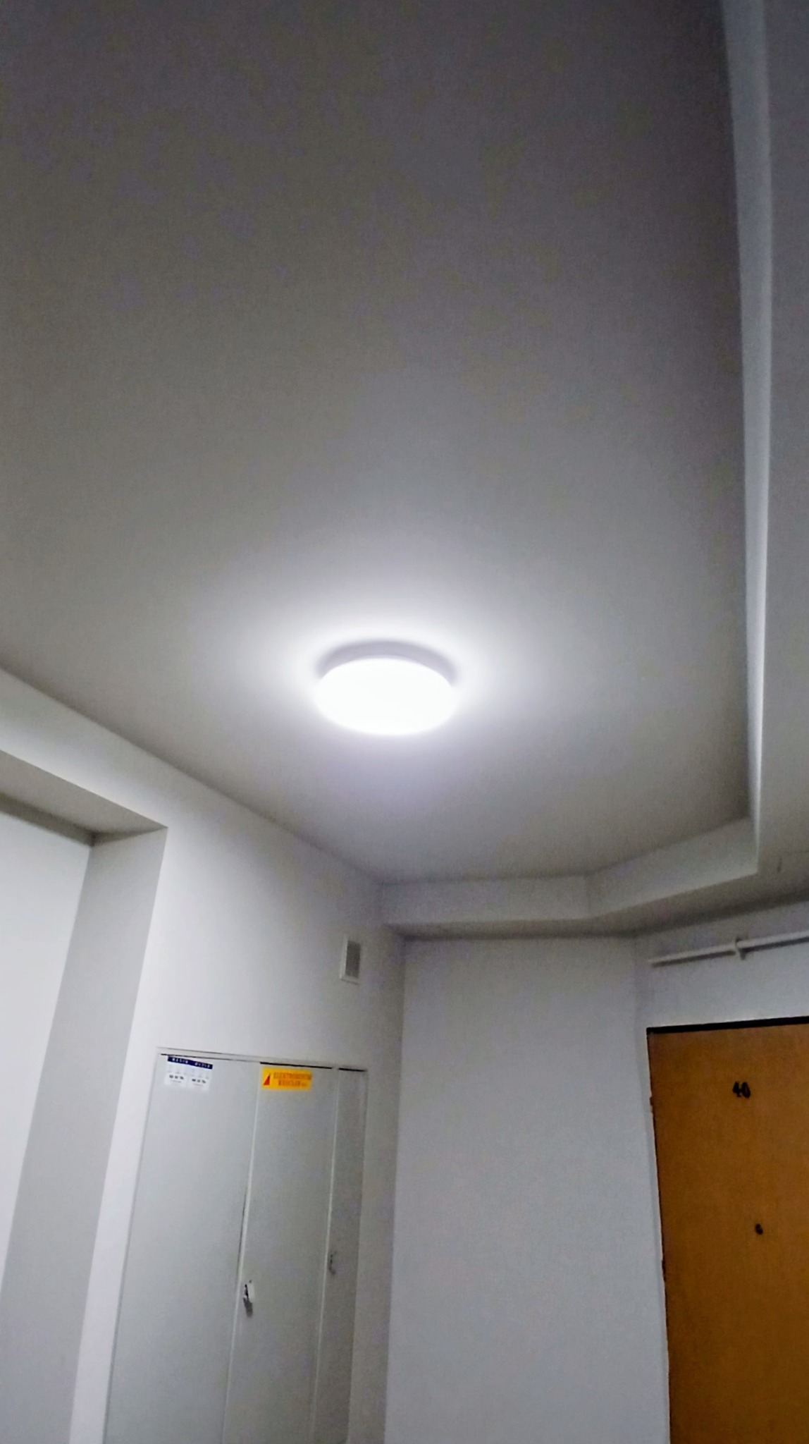 Białe wnętrze korytarza z okrągłą lampą sufitową, widoczne drzwi z numerem 46 i szafka elektryczna. Świeżo pomalowane ściany w neutralnym odcieniu bieli.