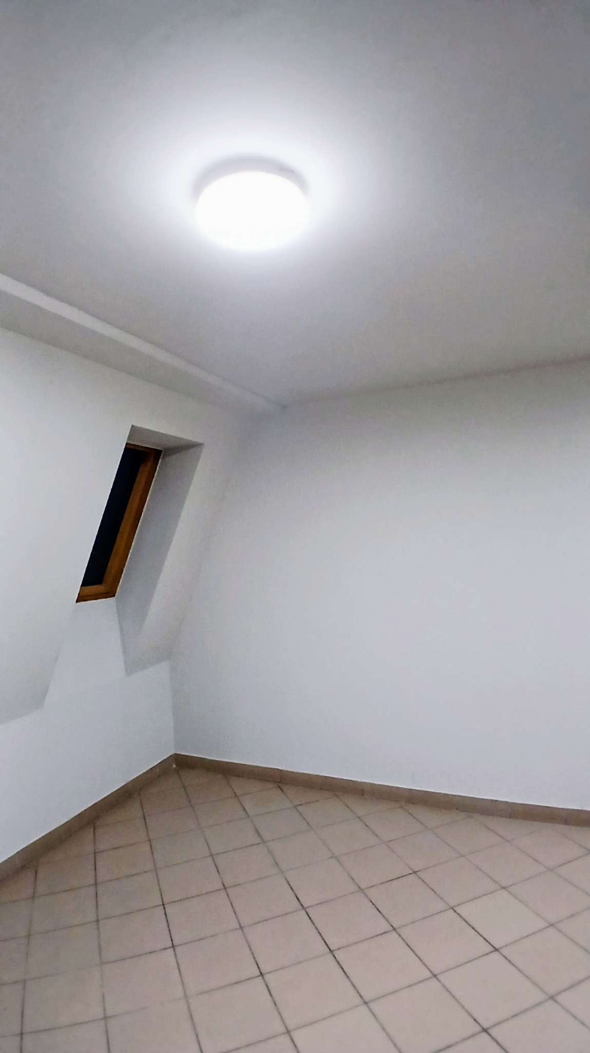 Wnętrze pokoju po remoncie w Rościsławicach. Białe ściany, beżowa podłoga z płytek, skośne okno dachowe i okrągła lampa sufitowa.