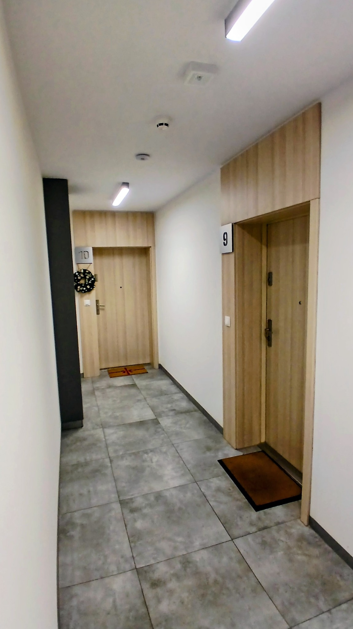 Korytarz w bloku z szarymi płytkami podłogowymi i drewnopodobnymi drzwiami do mieszkań nr 9 i 10, z wycieraczkami i wieńcem na drzwiach.