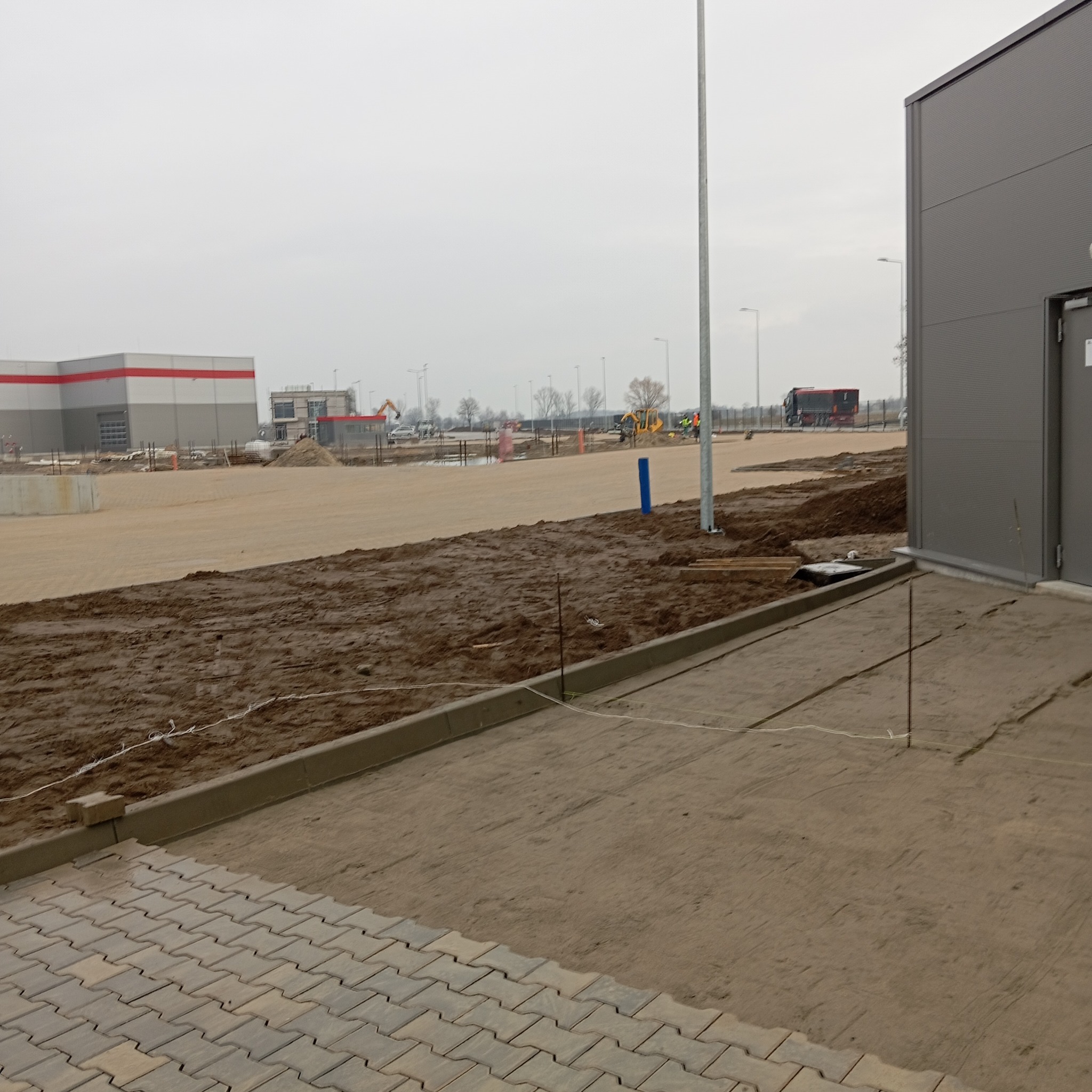 Fragment placu budowy z częściowo ułożoną kostką brukową, widoczne przygotowane podłoże, obrzeża betonowe, w tle budynki przemysłowe i maszyny budowlane, pochmurny dzień.