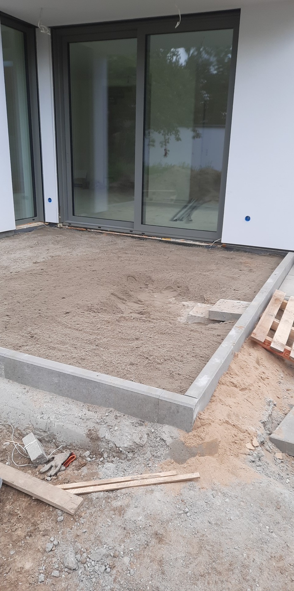 Przygotowanie podłoża pod kostkę brukową: wyrównany piasek w obramowaniu z betonowych obrzeży, widoczne narzędzia pracy i paleta w tle, ściana budynku z oknami przesuwnymi.