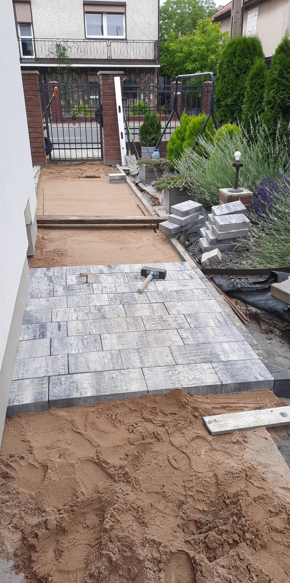 Fragment układanej ścieżki z szarych, prostokątnych płyt betonowych, widoczny młotek brukarski, poziomica i stos kostki brukowej w tle, piasek podsypkowy.