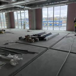 Andrii Pominchuk - Wnętrze w stanie surowym z widoczną instalacją wentylacyjną pod sufitem, przygotowane podłoże pod instalacje elektryczne i inne, duże okna z widokiem na budowę.