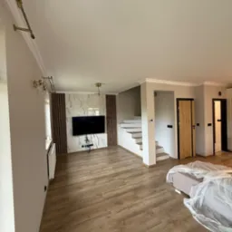 Jasne wnętrze po remoncie: drewniana podłoga, schody, dekoracyjna ściana z TV, listwy przysufitowe, oświetlenie, drzwi wewnętrzne. Częściowo zakryte meble.