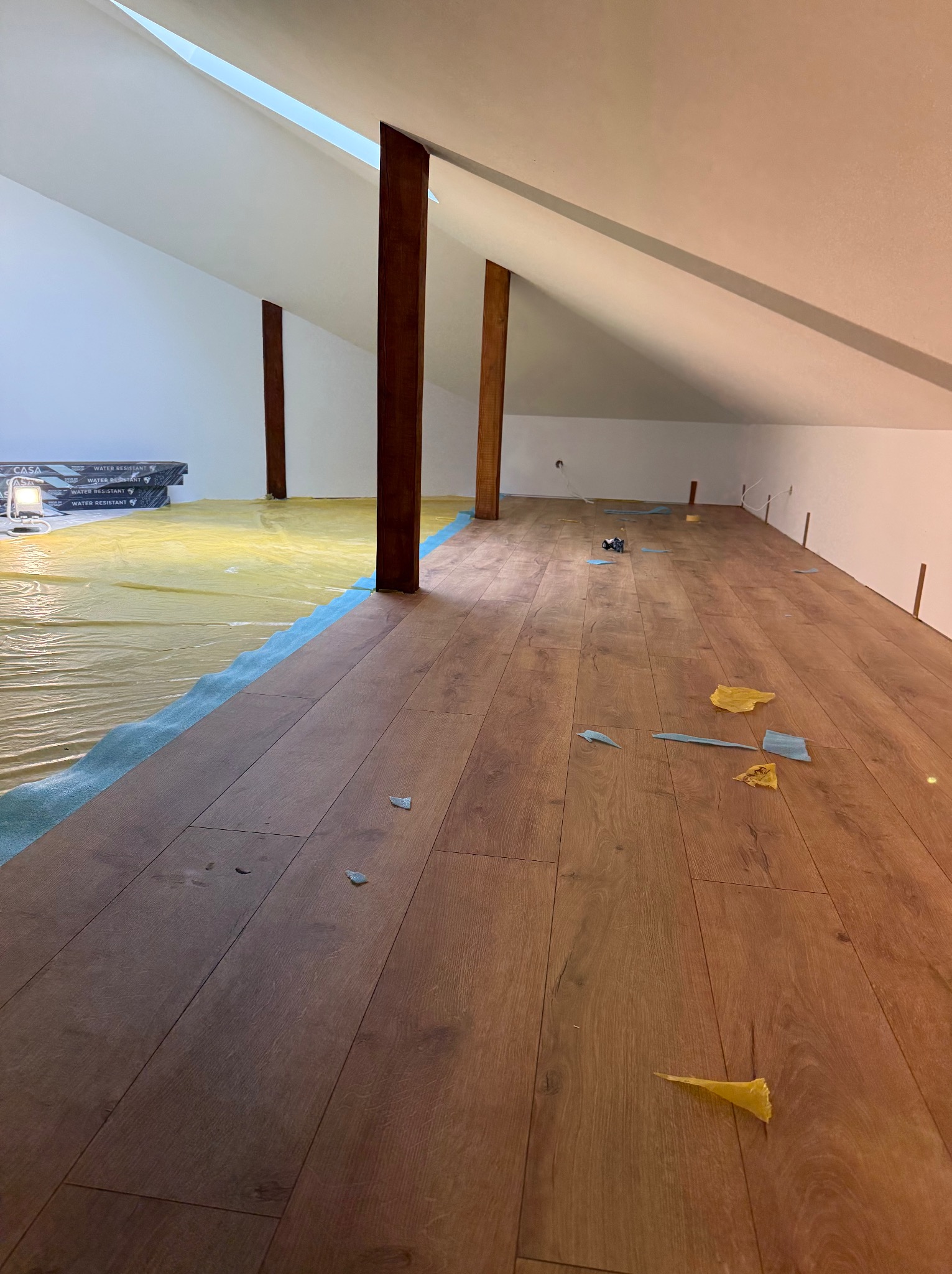 Podłoga z paneli w trakcie układania na poddaszu, widoczne folia ochronna, brązowe słupy konstrukcyjne i resztki po cięciu paneli.