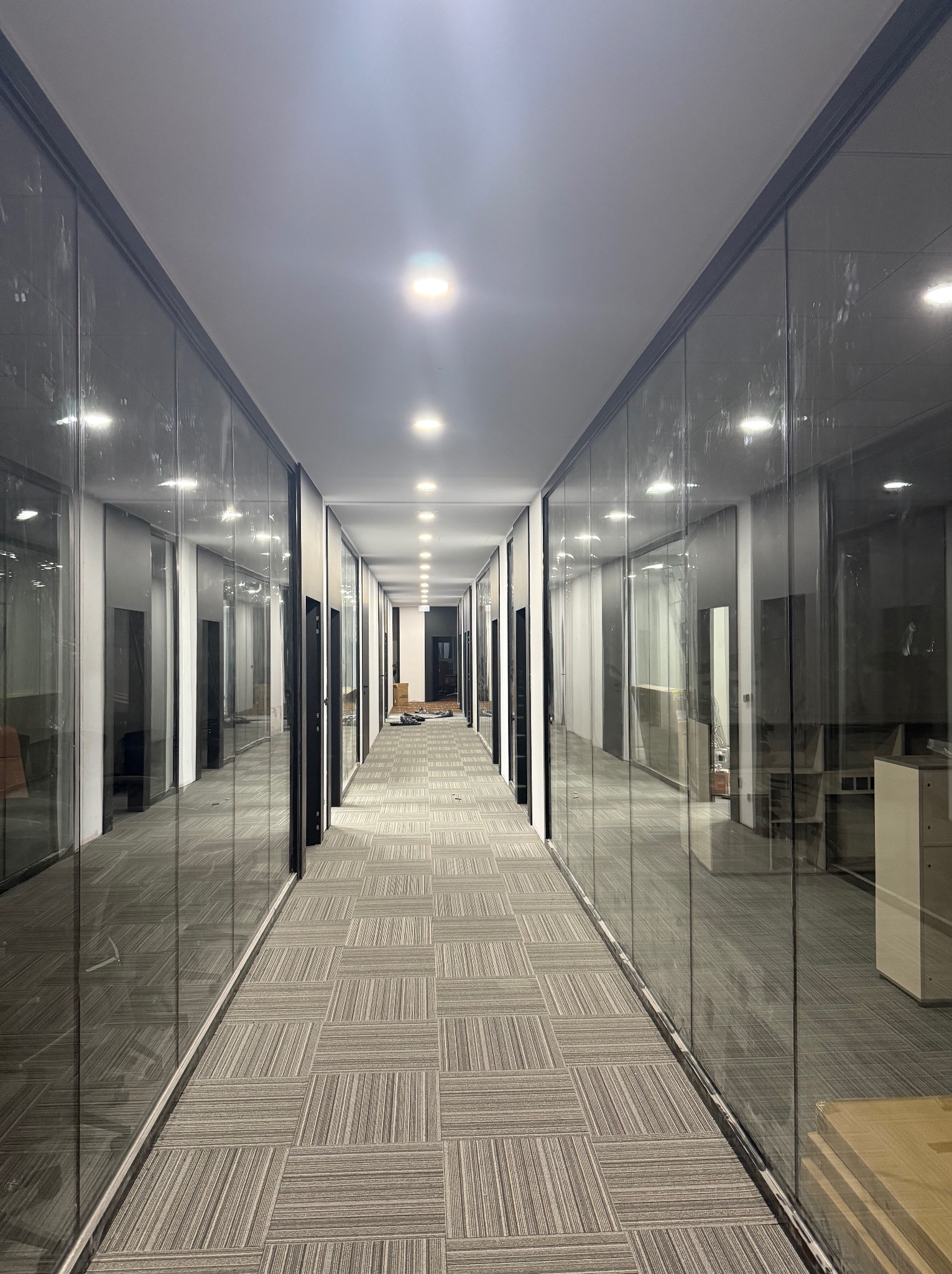 Długi korytarz biurowy z przeszklonymi ścianami działowymi, wzorzysta wykładzina podłogowa, widoczne elementy instalacji elektrycznej pod sufitem, częściowo umeblowane pomieszczenia.