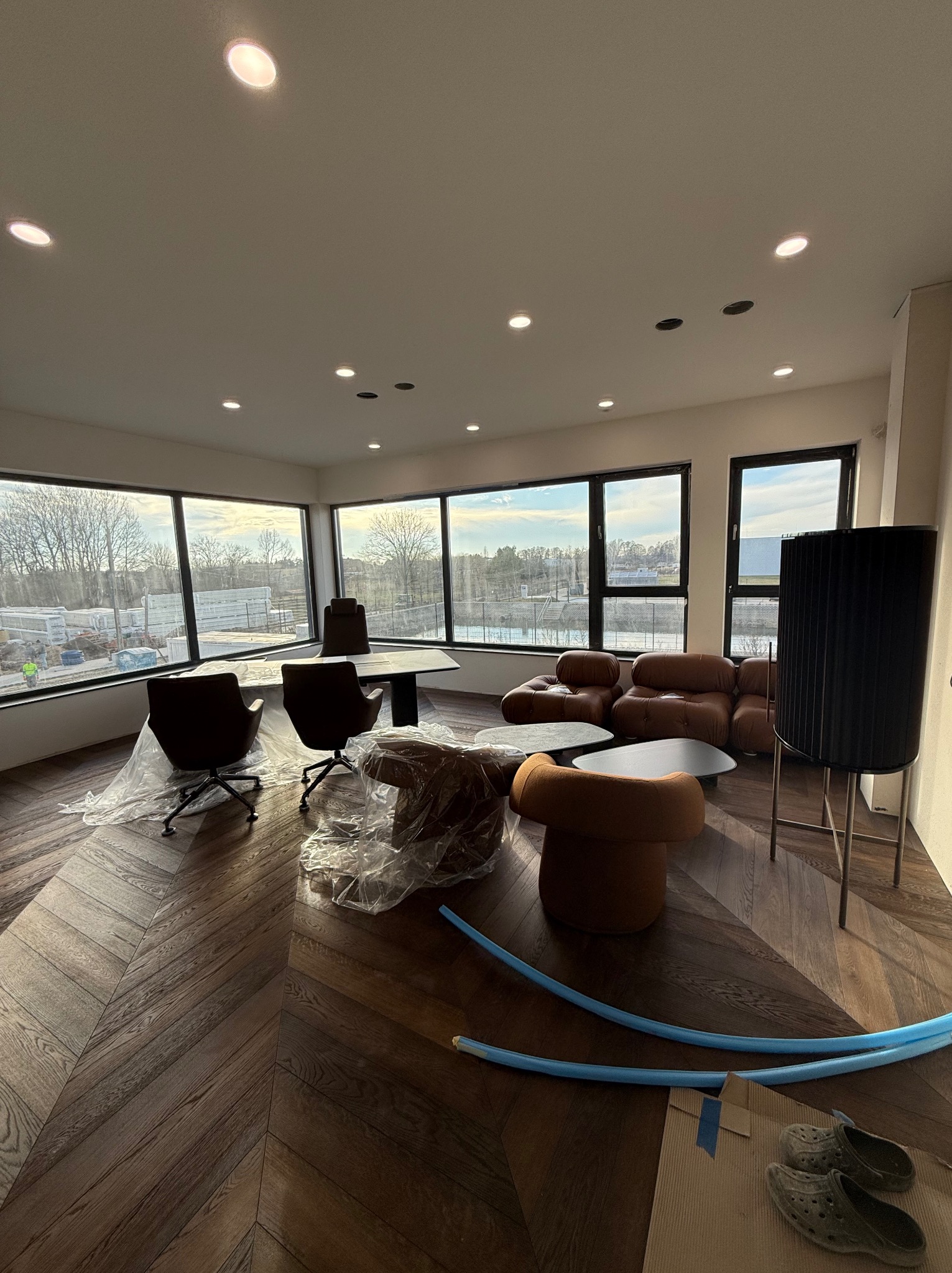 Wnętrze biura w trakcie wykańczania: widoczne nowe oświetlenie sufitowe typu downlight, meble zabezpieczone folią, parkiet w jodełkę, niebieskie rurki peszel i robocze obuwie na podłodze.