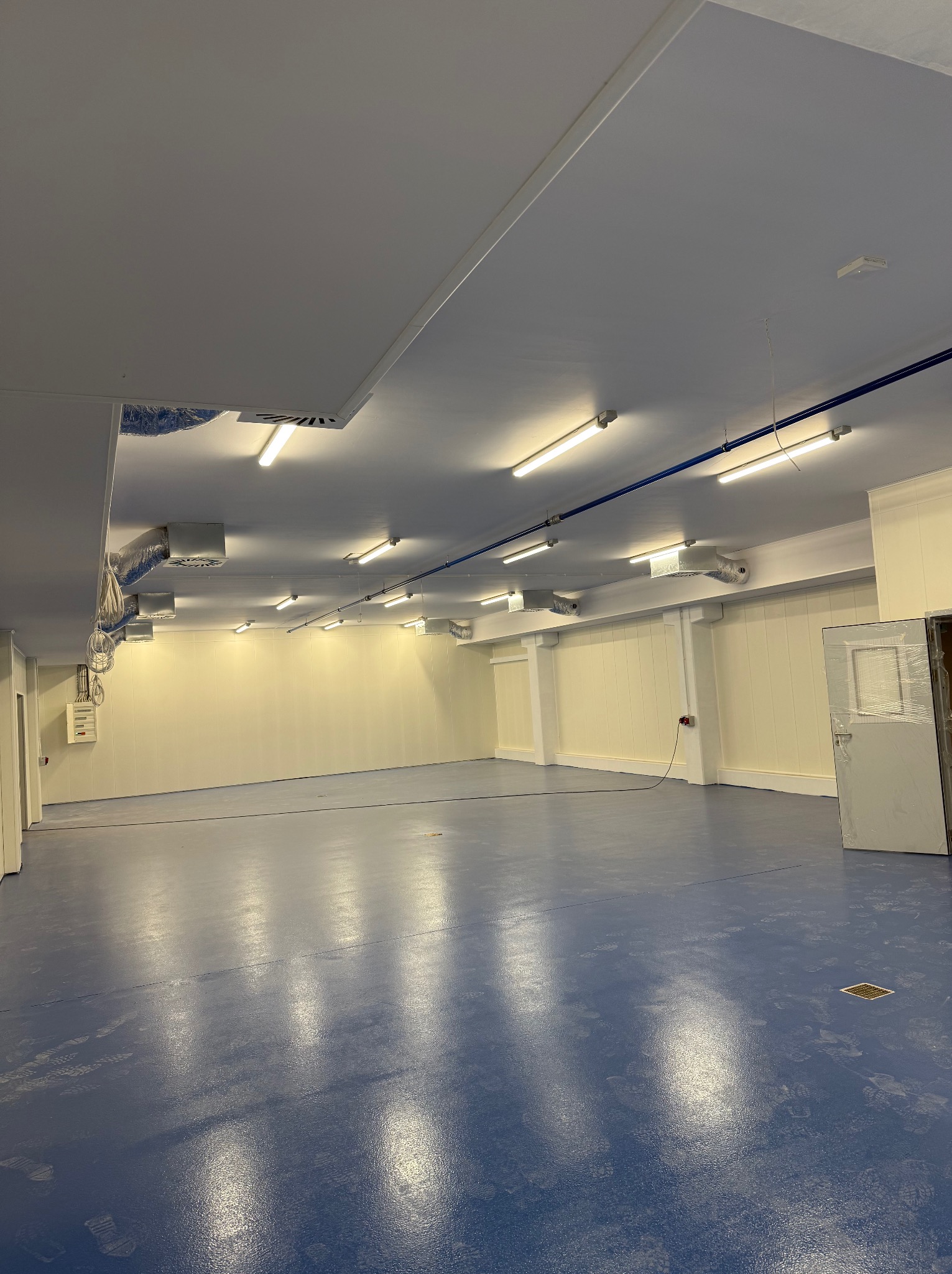 Wnętrze hali produkcyjnej po remoncie, z widocznym oświetleniem liniowym LED, niebieską posadzką żywiczną i instalacją wentylacyjną pod sufitem.