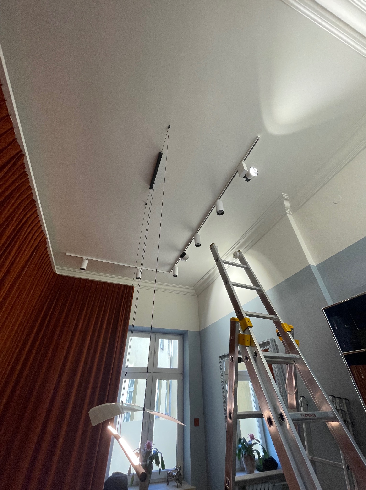 Widok z dołu na sufit z zamontowanym oświetleniem szynowym i lampą wiszącą, z rozstawioną drabiną aluminiową, zasłonięte okno z boku.