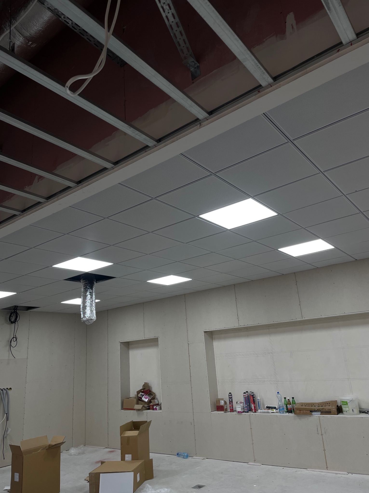 Sufit podwieszany z zamontowanymi panelami oświetleniowymi, widoczne prace wykończeniowe ścian i instalacja wentylacyjna z folii aluminiowej.