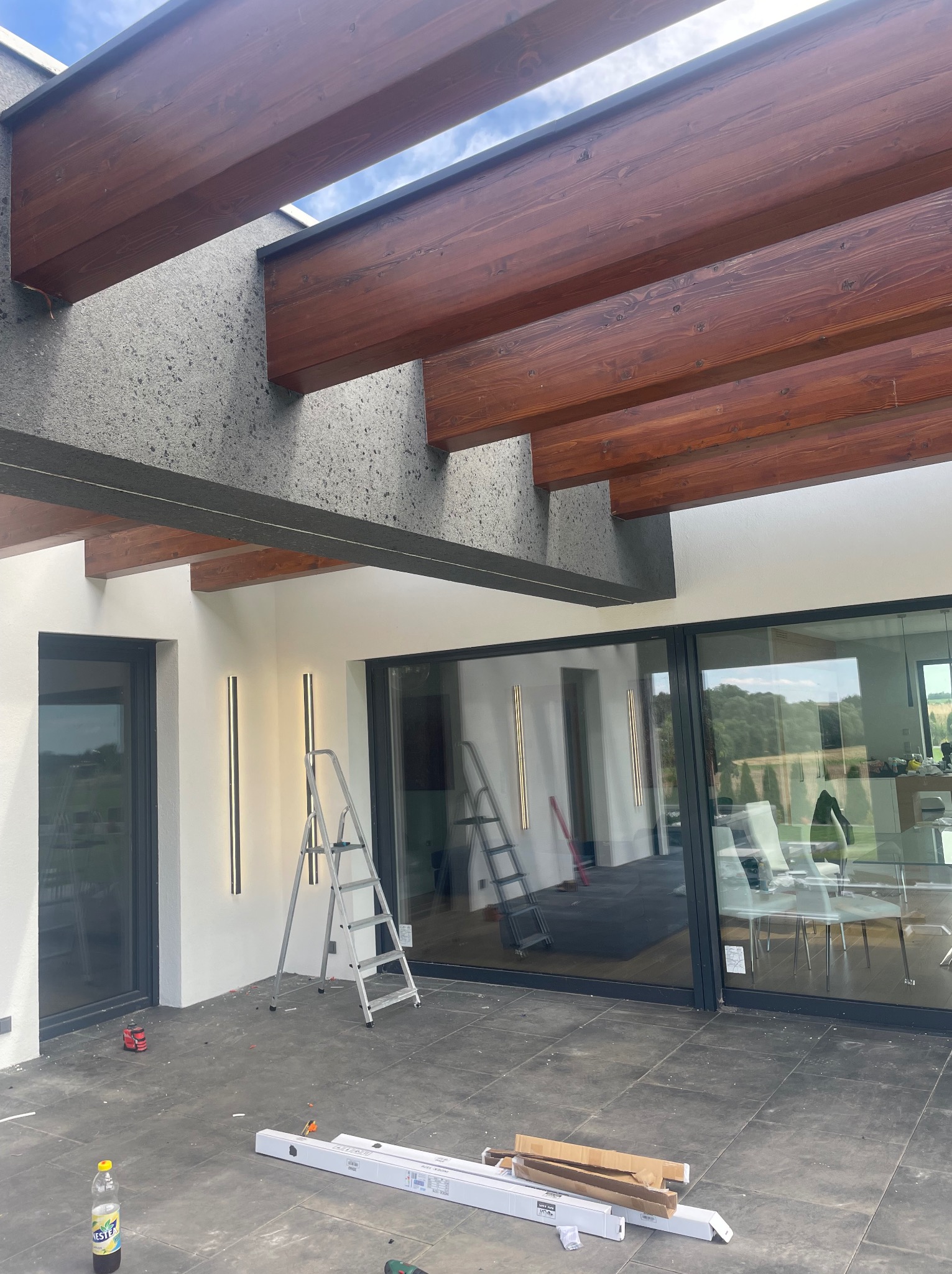 Montaż pionowych lamp LED na elewacji domu z użyciem drabiny, widoczne drewniane belki zadaszenia i narzędzia pracy.