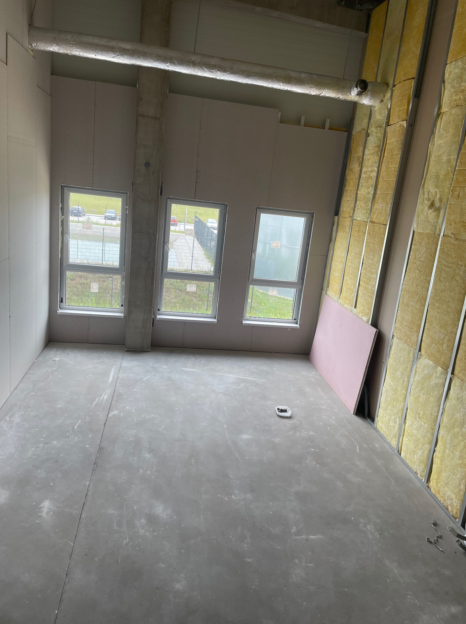 Wnętrze w trakcie wykańczania w Warszawie: ściany z izolacją z wełny mineralnej, surowy beton na podłodze, trzy okna wpuszczające światło dzienne, widoczne przewody wentylacyjne pod sufitem.