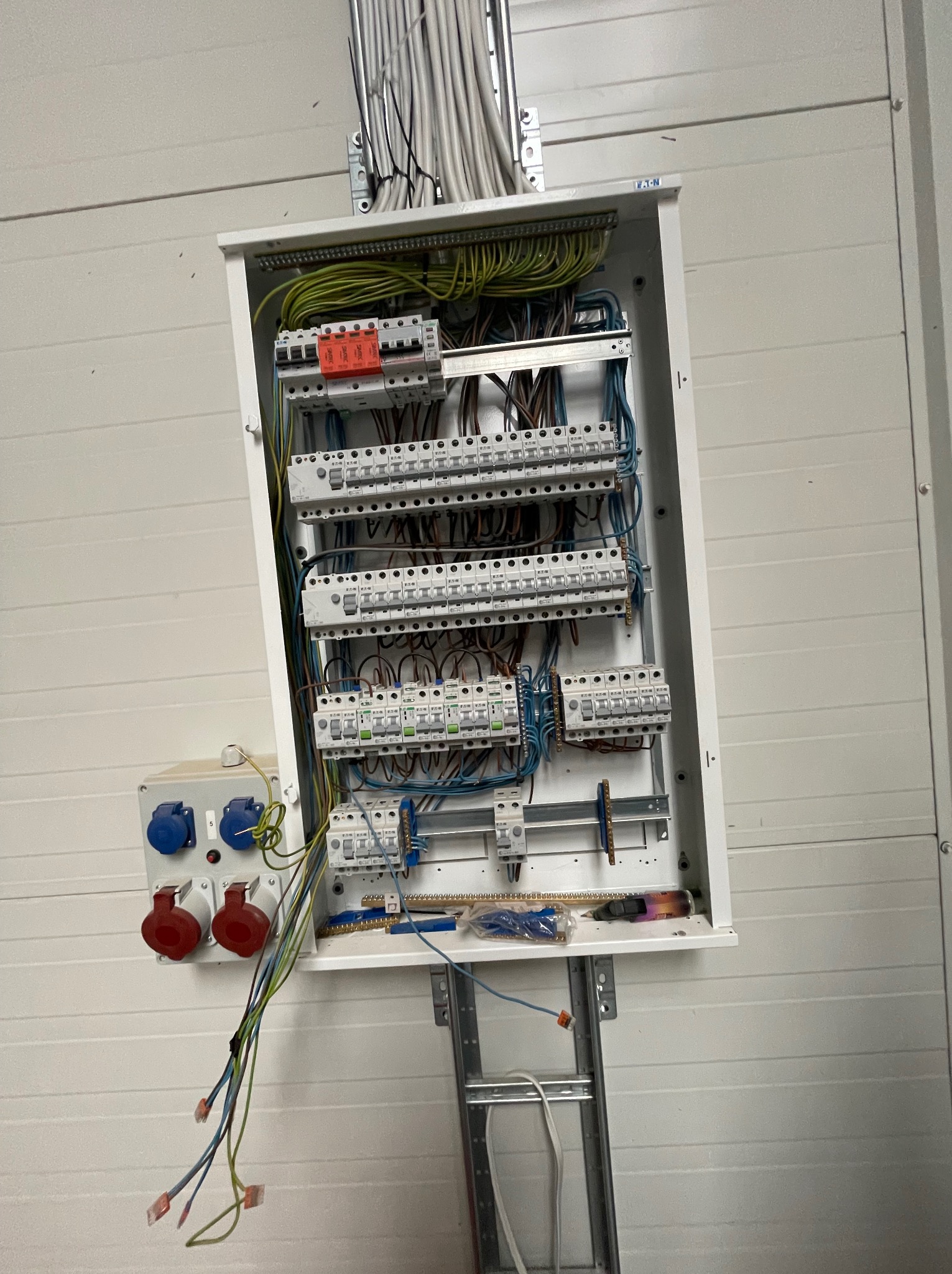 Otwarta skrzynka elektryczna firmy Eaton z licznymi zabezpieczeniami i przewodami, zamontowana na białej ścianie z widocznymi gniazdami siłowymi, na metalowej drabince.