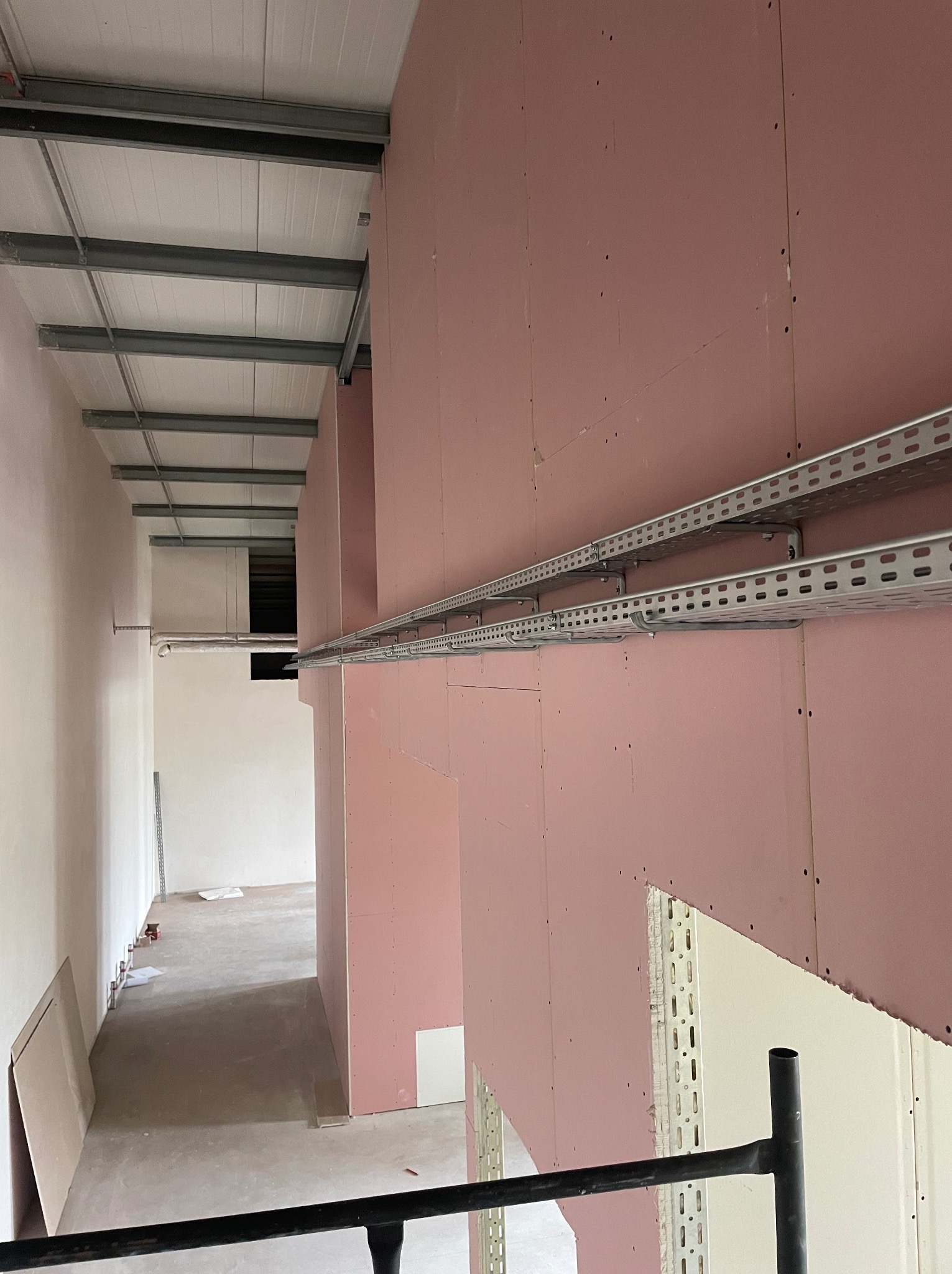 Wnętrze w trakcie wykańczania: różowe płyty gipsowo-kartonowe na ścianach, metalowe korytka kablowe zamontowane pod sufitem, widok z góry na surową podłogę i dalsze pomieszczenia.