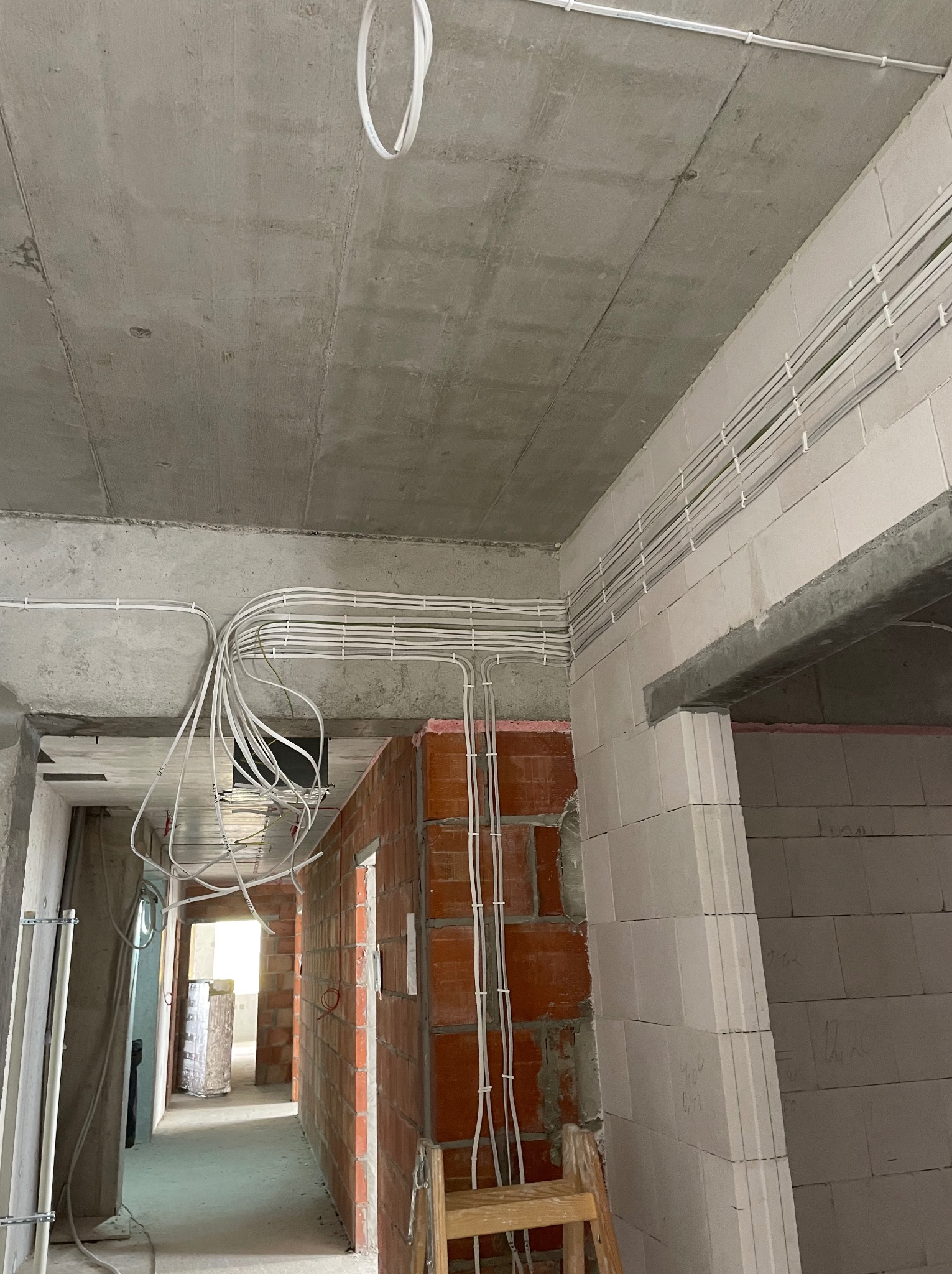 Wnętrze w stanie surowym z widoczną instalacją elektryczną: białe przewody prowadzone po suficie i ścianach, mocowane uchwytami, z widocznym rozdzieleniem przewodów i przejściem przez ściany...