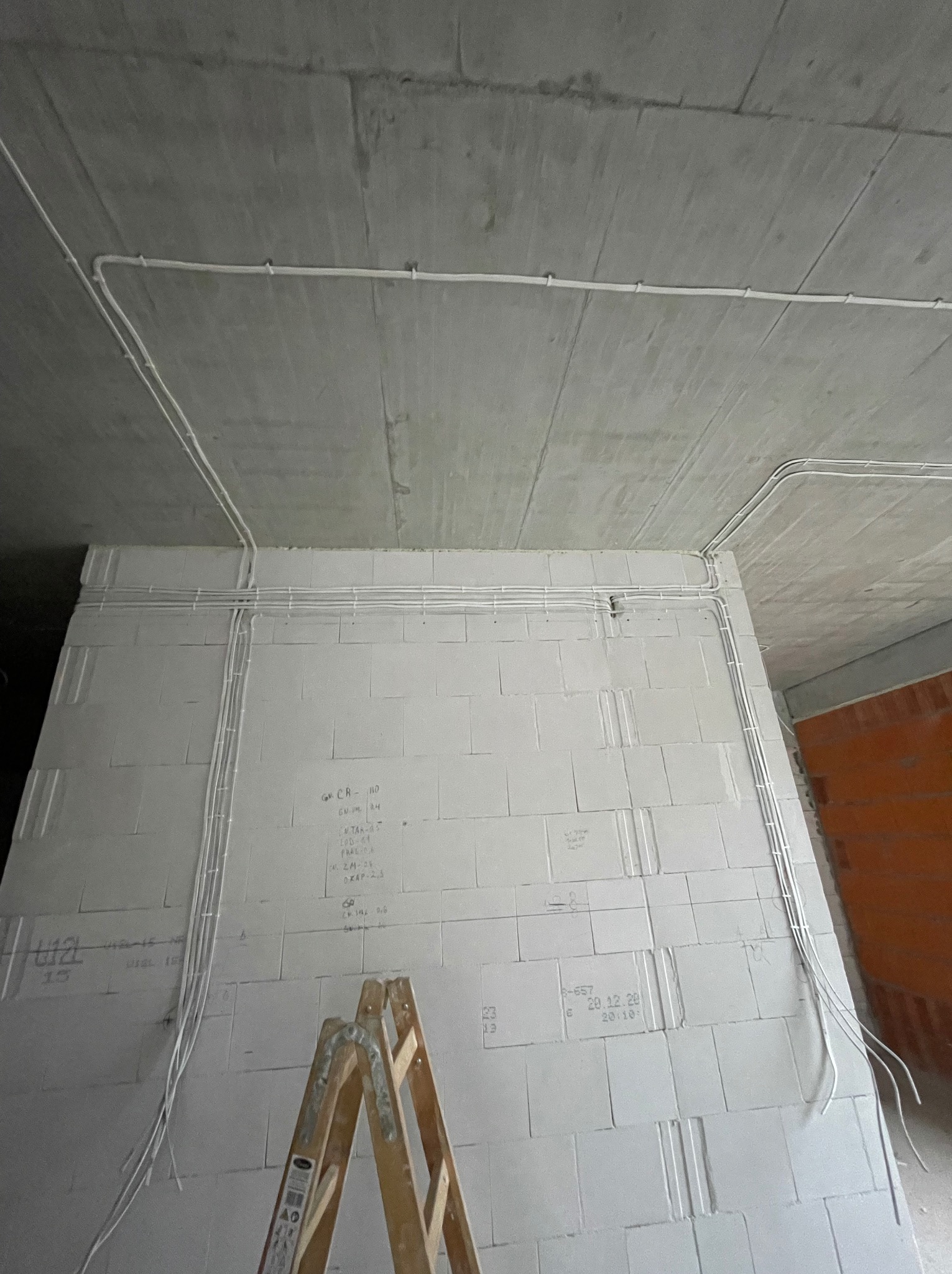 Wewnętrzne rozprowadzenie przewodów elektrycznych po ścianach z betonu komórkowego w surowym stanie, widoczne napisy techniczne i drabina przystawna