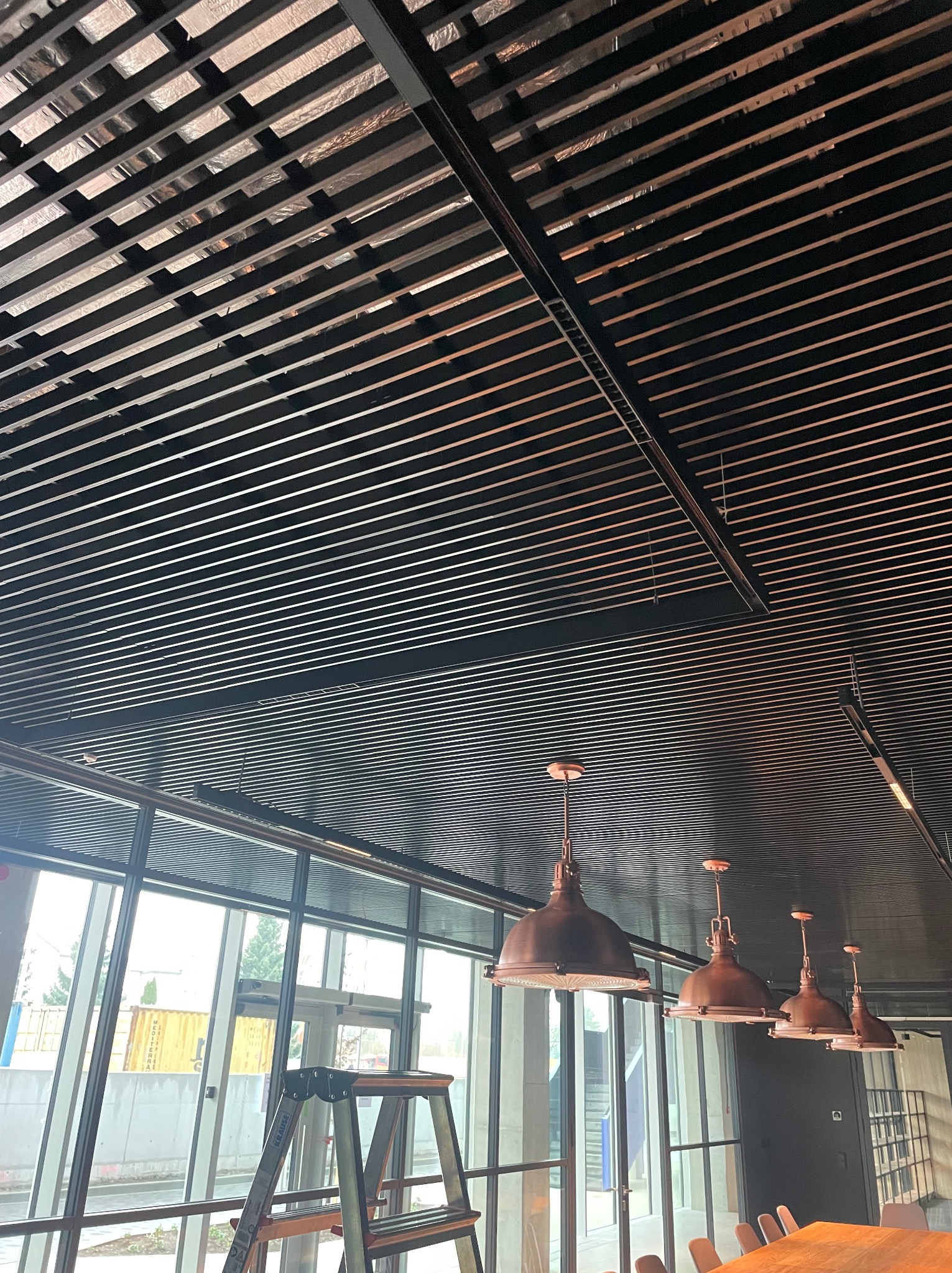 Widok na sufit z drewnianą kratownicą i rzędem miedzianych lamp wiszących, drabina oparta o okno w tle.