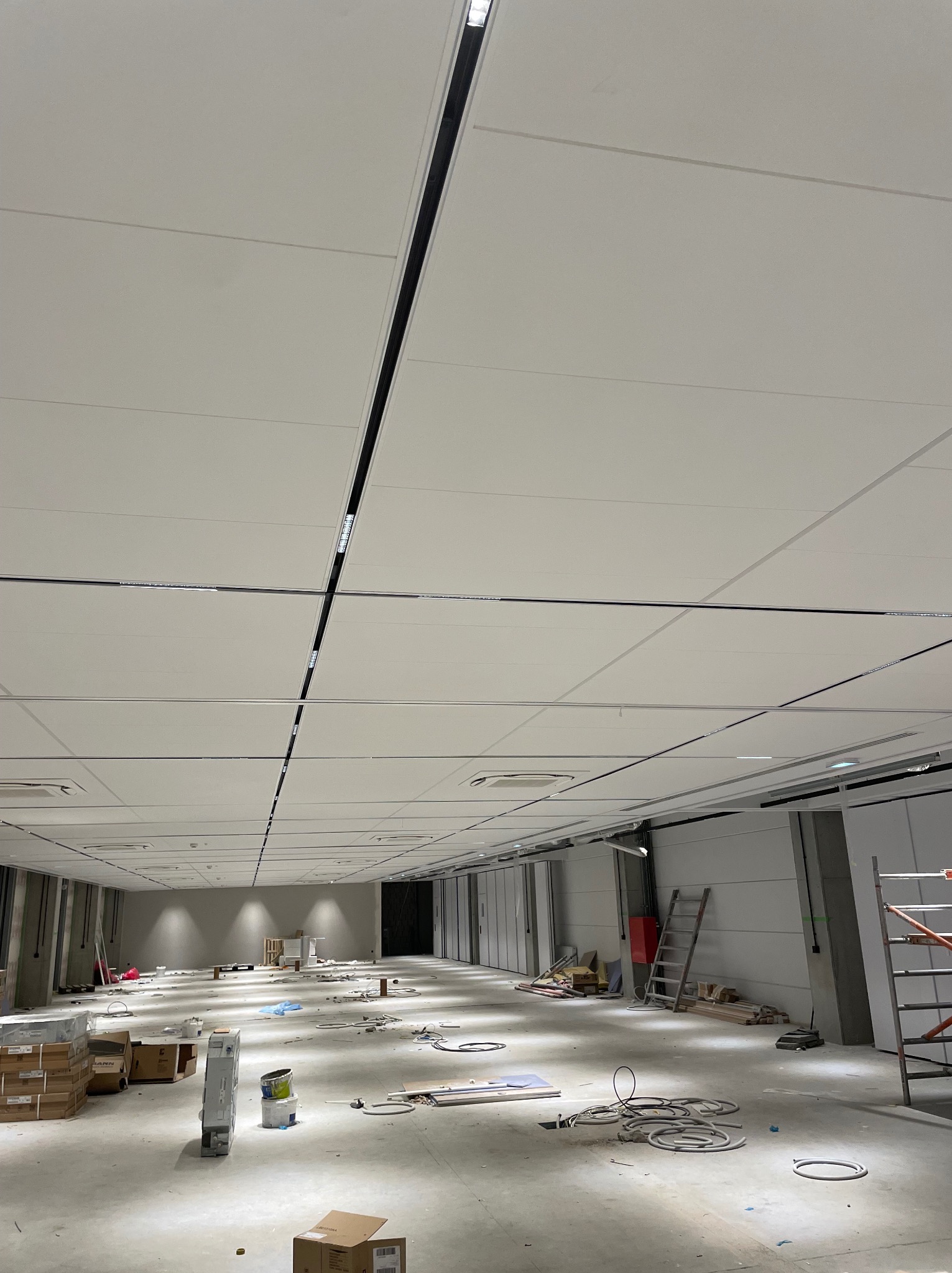 Wnętrze w trakcie prac wykończeniowych, widoczne kable elektryczne na podłodze, drabina oparta o ścianę i podwieszony sufit z zamontowanym oświetleniem liniowym.