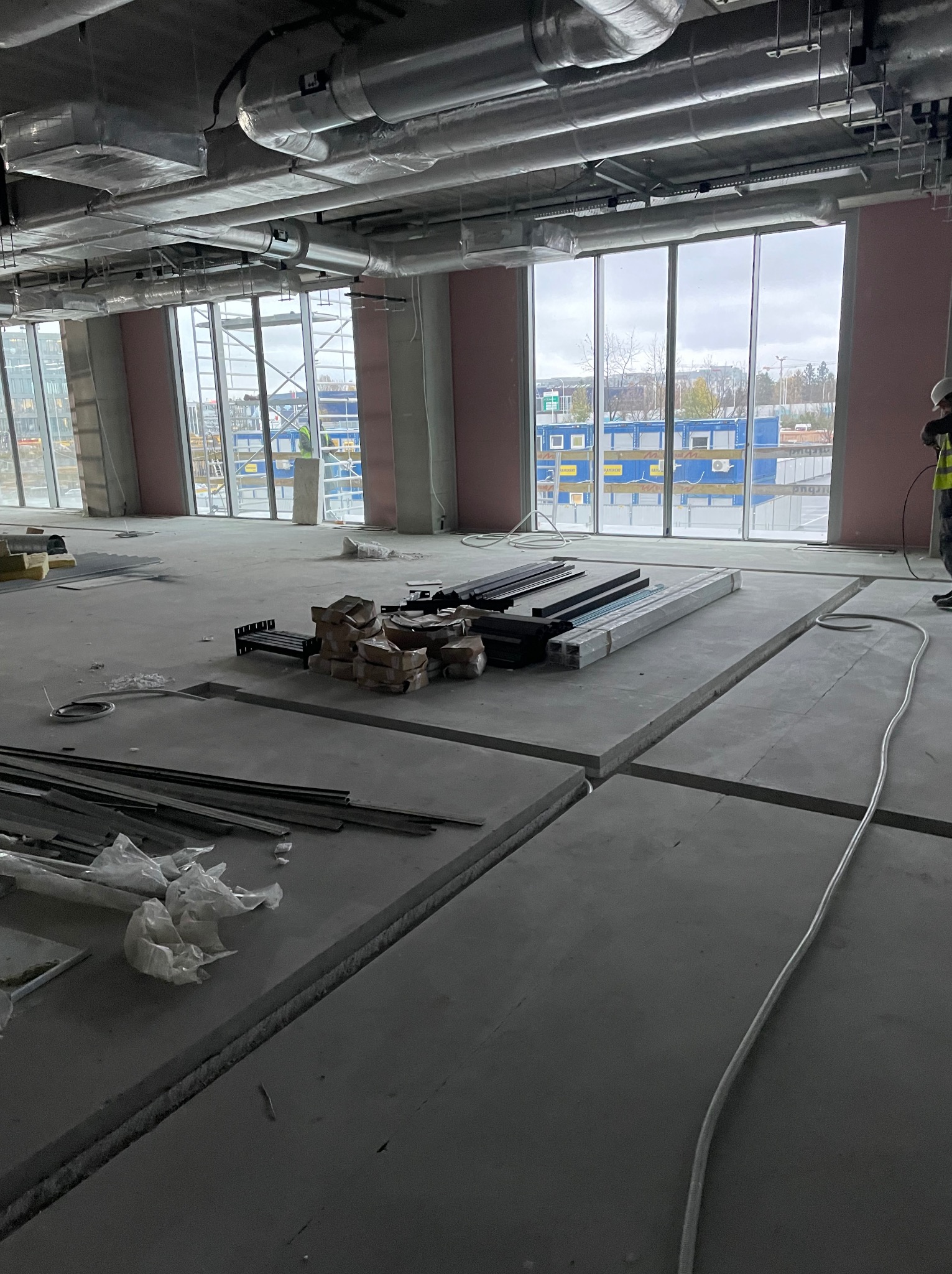 Wnętrze w stanie surowym z widoczną instalacją wentylacyjną pod sufitem, przygotowane podłoże pod instalacje elektryczne i inne, duże okna z widokiem na budowę.