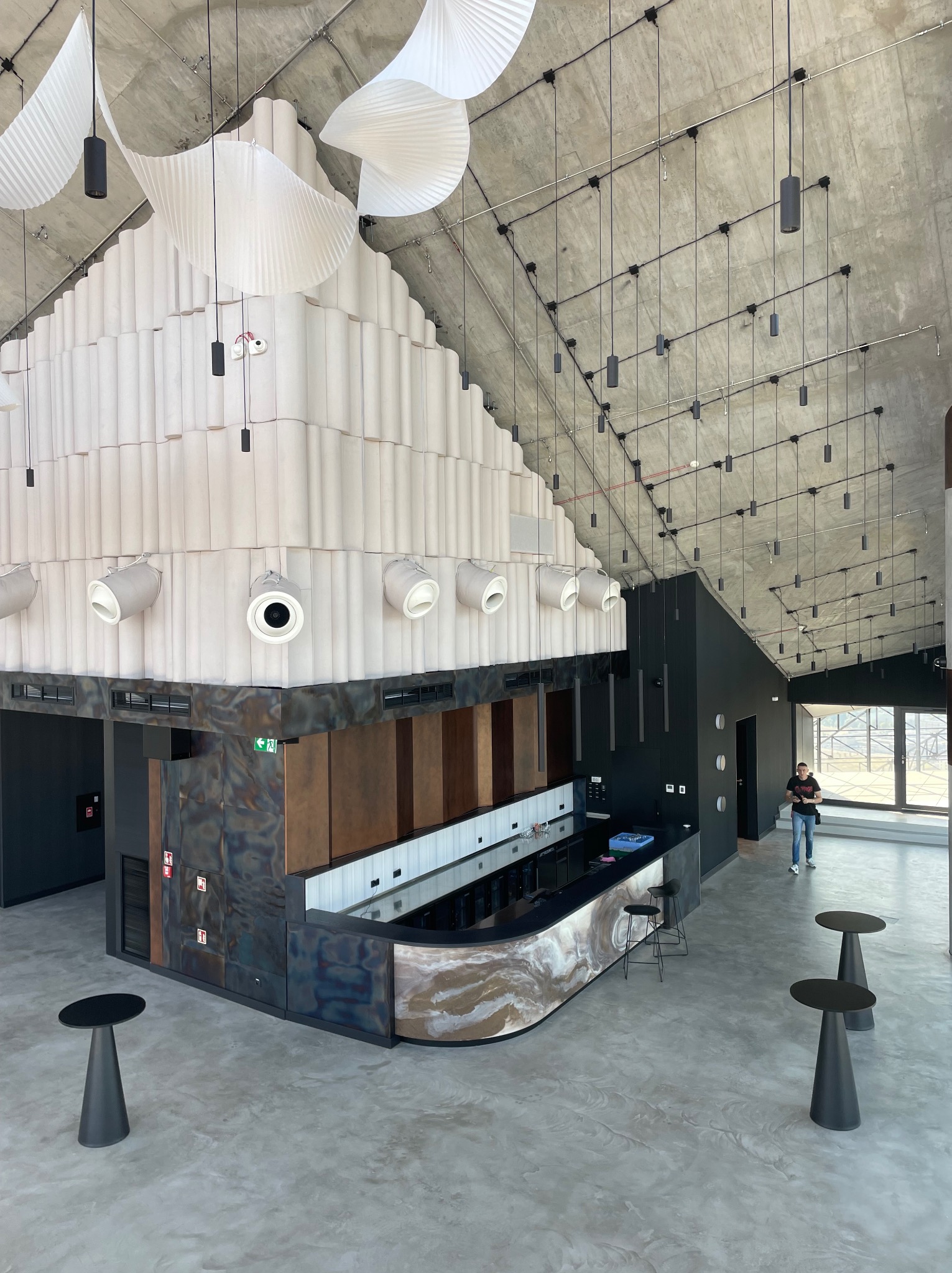 Wnętrze nowoczesnego baru z surowym betonowym sufitem, podwieszanymi lampami, dekoracyjną ścianą z podświetleniem i osobą w tle.