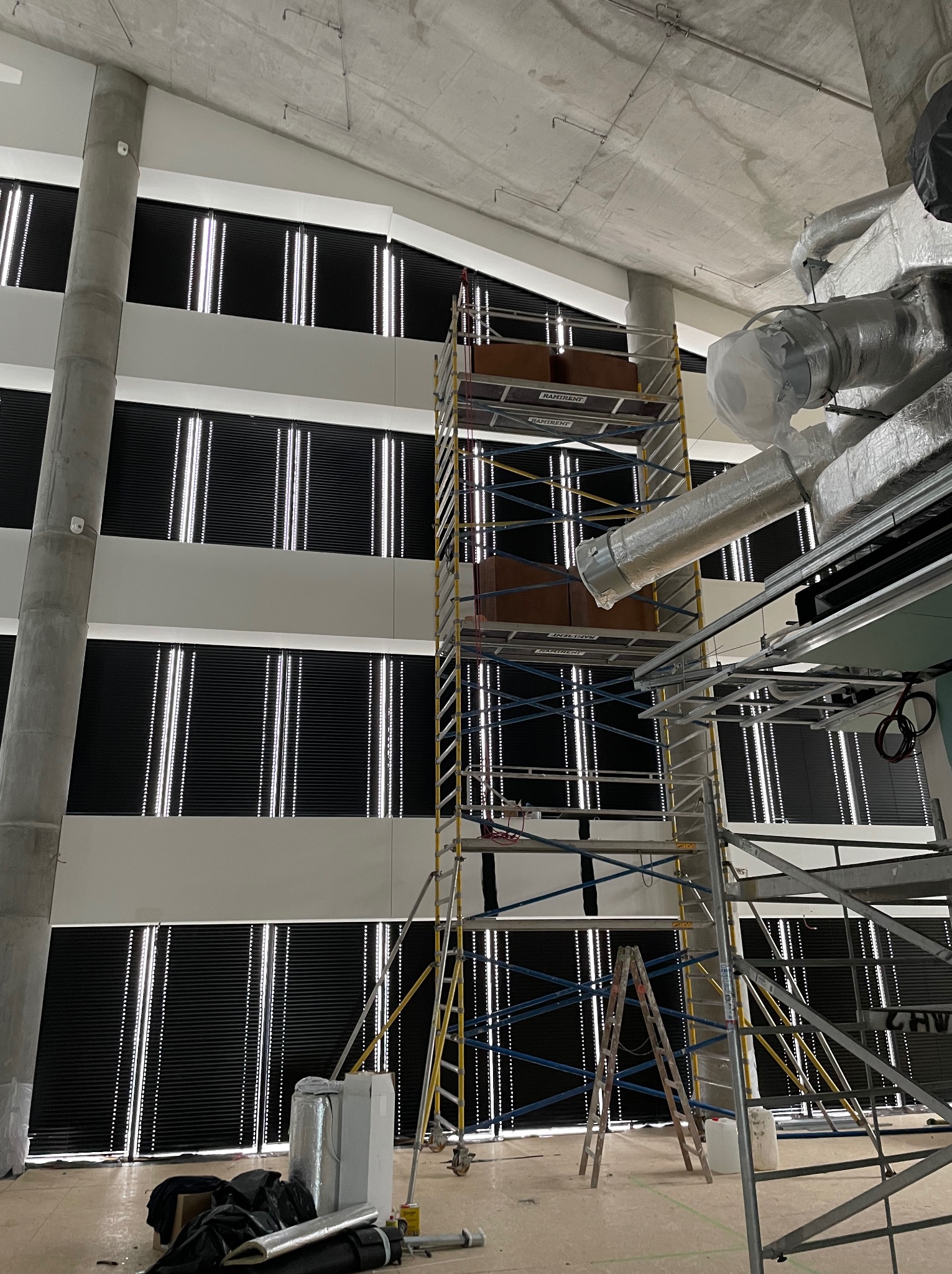 Wysokie rusztowanie na tle ściany z zamontowanymi, podświetlanymi żaluzjami w surowym wnętrzu budynku w trakcie prac instalacyjnych.