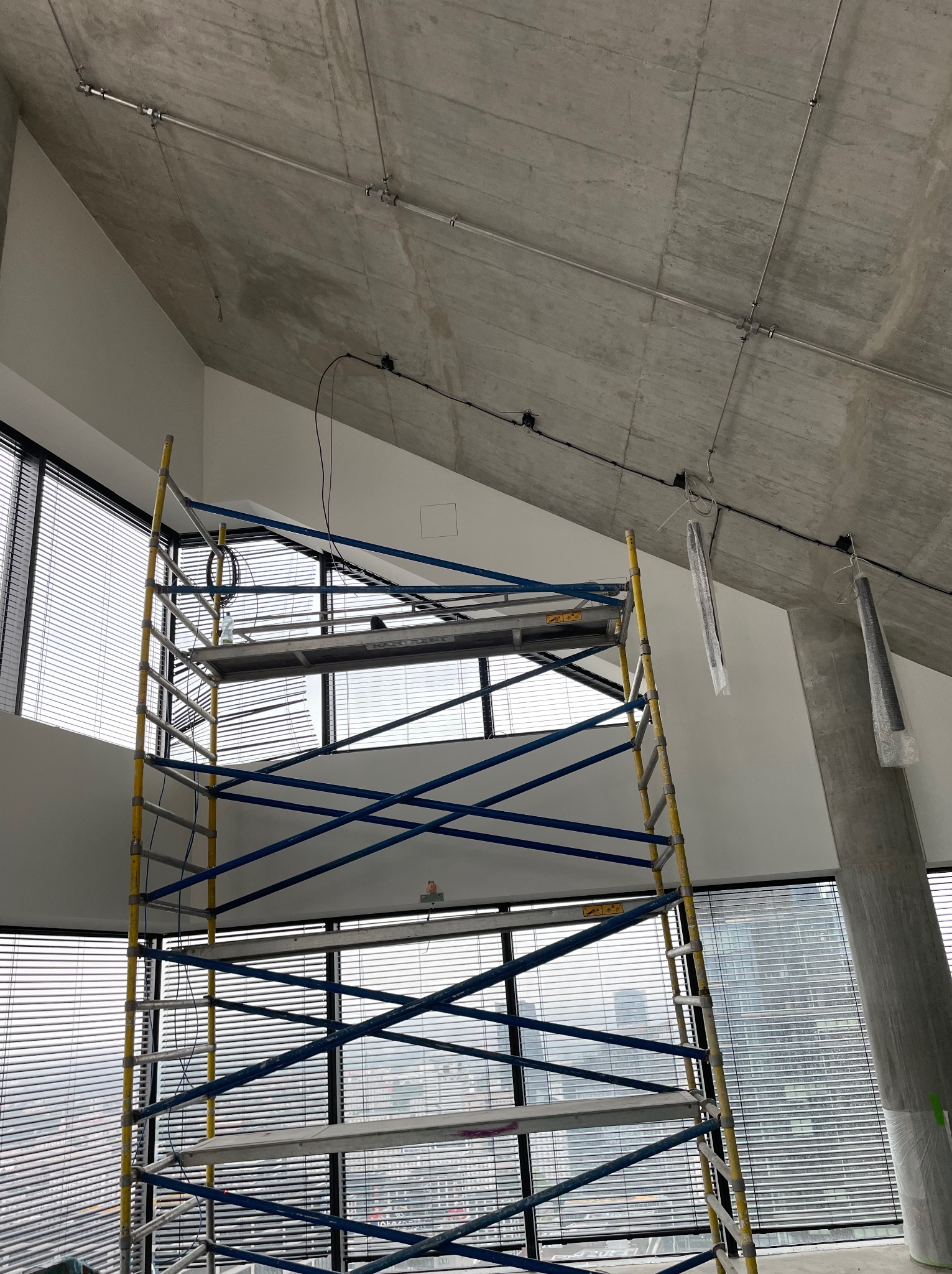 Wysokie rusztowanie na tle okna z żaluzjami i surowego betonowego sufitu z widocznymi rurami instalacji elektrycznej oraz wiszącymi lampami w folii ochronnej.
