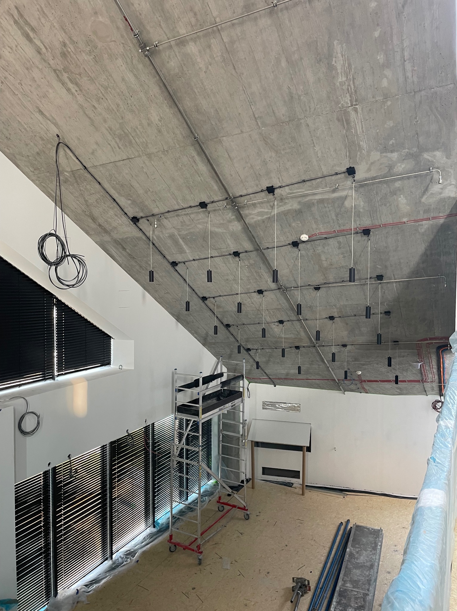 Wysoki sufit z betonu architektonicznego, na którym zamontowano system szynoprzewodów z czarnymi, cylindrycznymi lampami wiszącymi. Widoczny rusztowanie i zwisające kable elektryczne przy oknie...