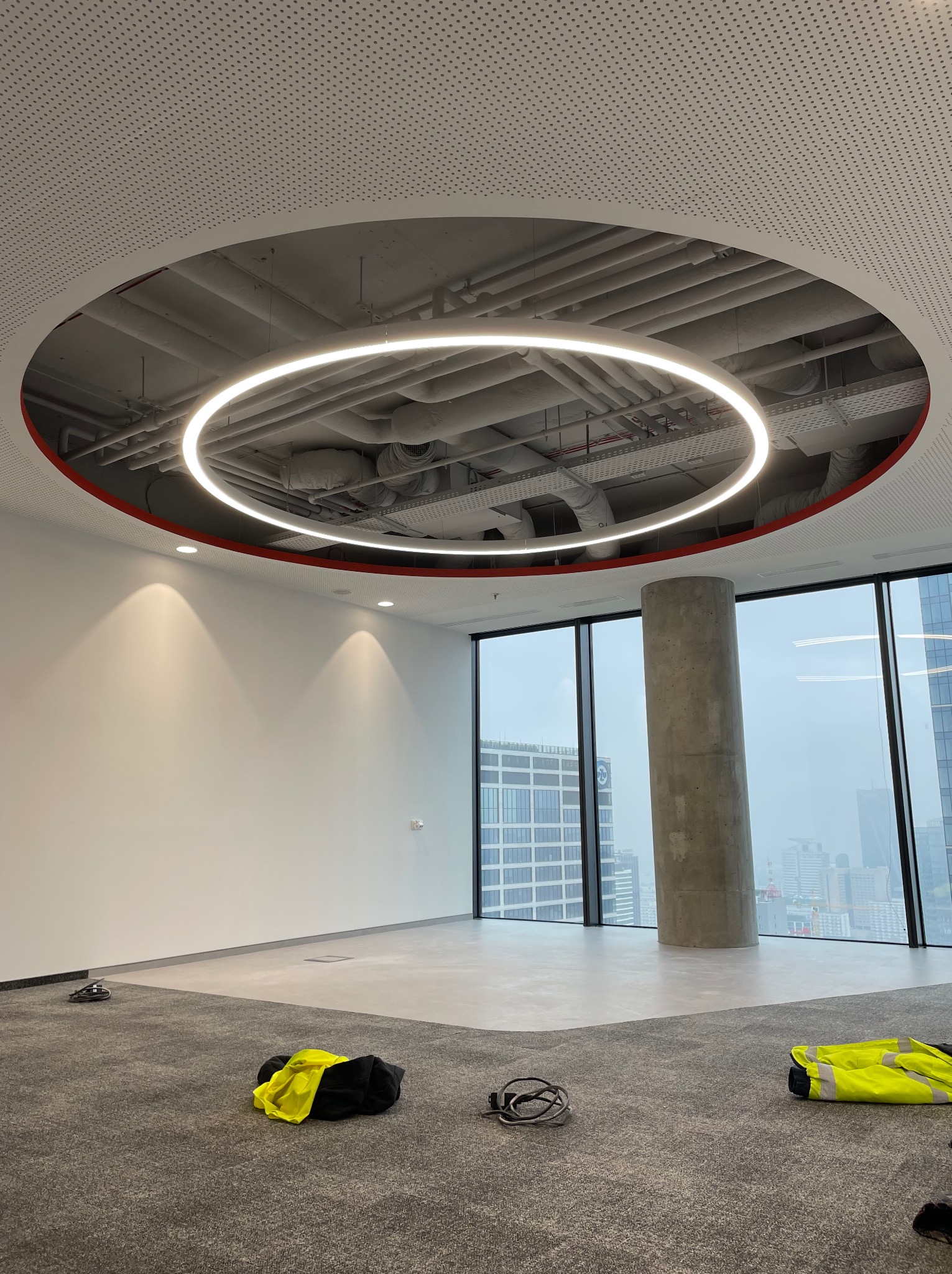 Wnętrze biurowe z okrągłą lampą LED pod sufitem, widoczne instalacje pod sufitem, kolumna betonowa przy oknie, oraz odzież robocza na podłodze.