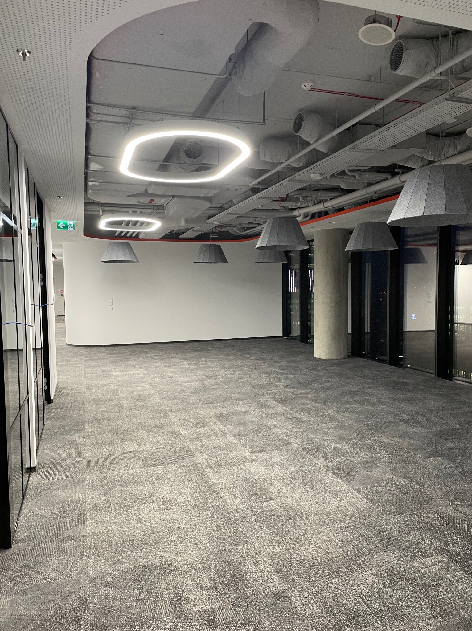 Wnętrze biurowe z szarą wykładziną dywanową, betonowym filarem i podwieszanymi lampami w kształcie stożków oraz kwadratowymi oprawami oświetleniowymi LED na suficie z widoczną instalacją wentylacyjną.