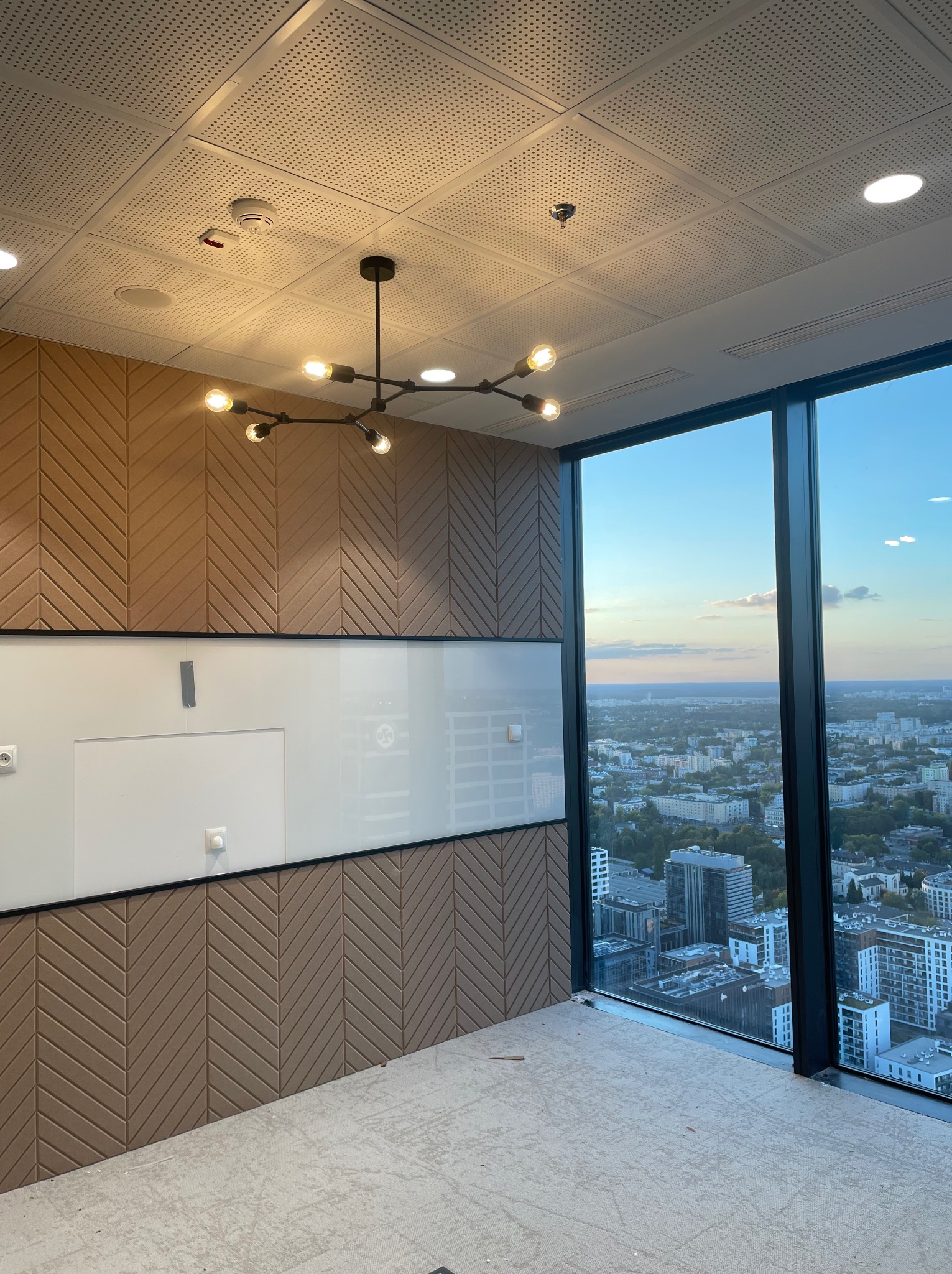 Nowoczesne biuro z panoramicznym widokiem na miasto, designerska lampa sufitowa z żarówkami Edisona, dekoracyjna ściana z panelami w jodełkę i biała tablica suchościeralna.