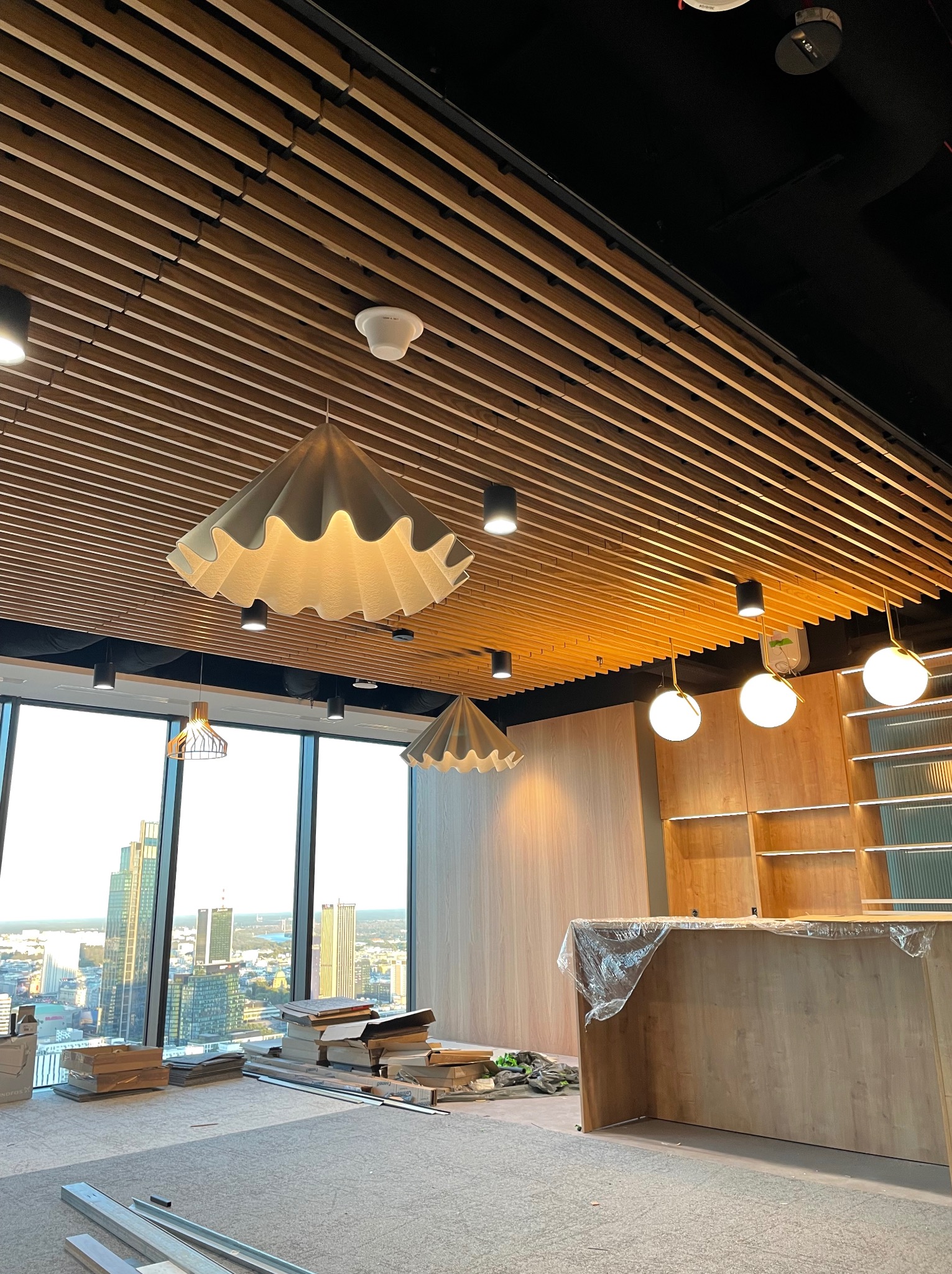 Wnętrze biurowe w trakcie wykańczania z drewnianym sufitem lamelowym, designerskimi lampami i widokiem na miasto przez panoramiczne okna. Na podłodze leżą materiały budowlane i narzędzia.