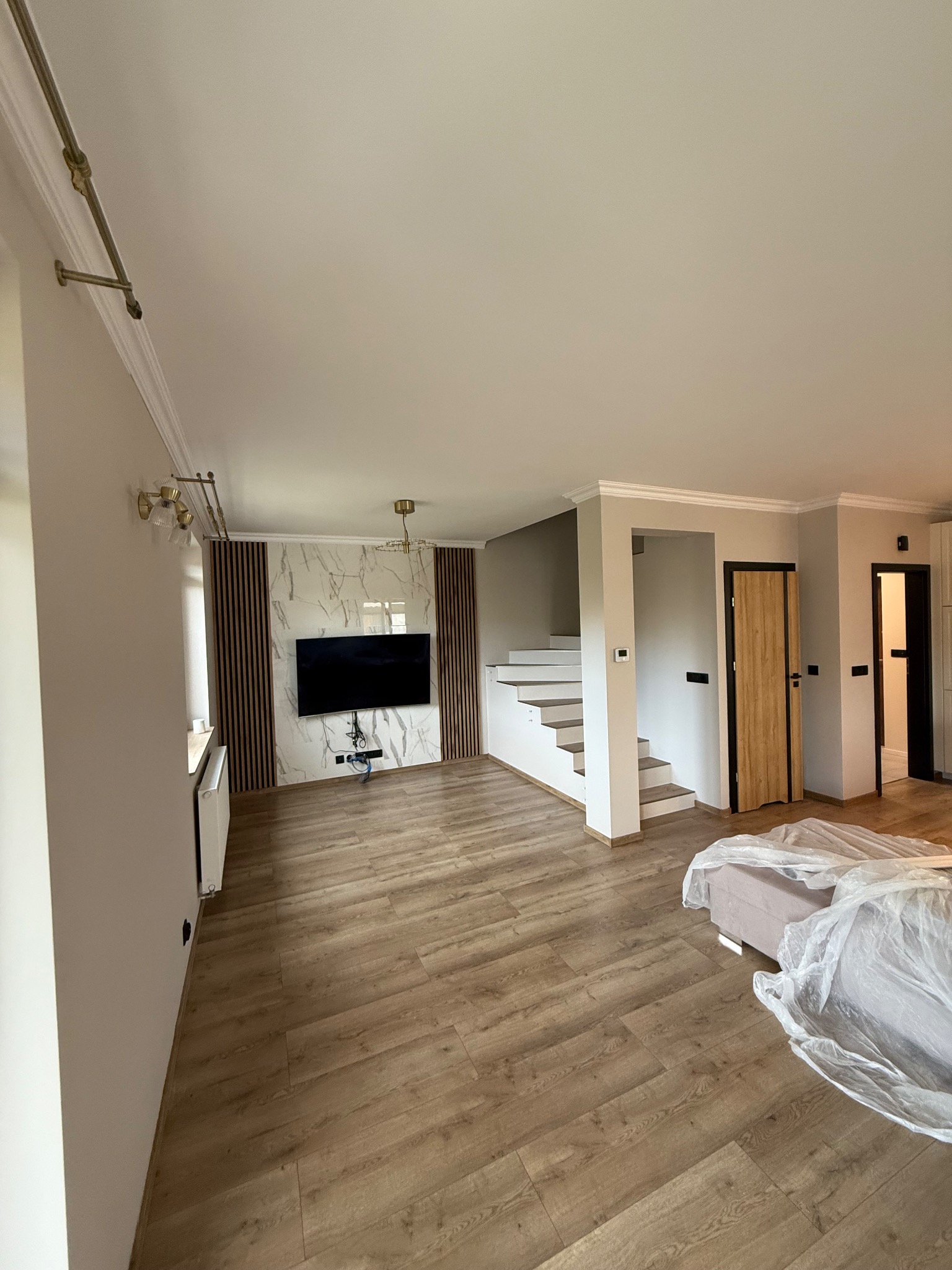 Jasne wnętrze po remoncie: drewniana podłoga, schody, dekoracyjna ściana z TV, listwy przysufitowe, oświetlenie, drzwi wewnętrzne. Częściowo zakryte meble.