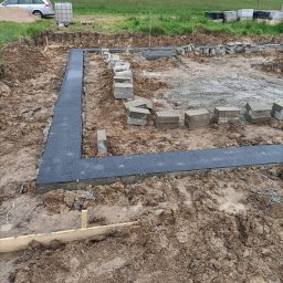 MAT-BUD Usługi ogólnobudowlane - Wykop pod fundamenty domu jednorodzinnego z częściowo ułożoną izolacją poziomą w postaci szarych płyt gumowych, obok ułożone bloczki fundamentowe, w tle samochód osobowy i budynki mieszkalne.