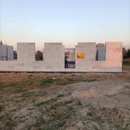 MAT-BUD Usługi ogólnobudowlane - Wznoszone ściany domu z białych bloczków, widoczny pracownik wewnątrz konstrukcji i żółta tablica informacyjna na ścianie.