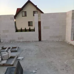 Wznoszenie ścian z białych bloczków betonu komórkowego na tle dwukondygnacyjnego domu z czerwonym dachem i zielonymi tujami w ogrodzie, widoczne drewniane palety z bloczkami i fragmenty gruzu...