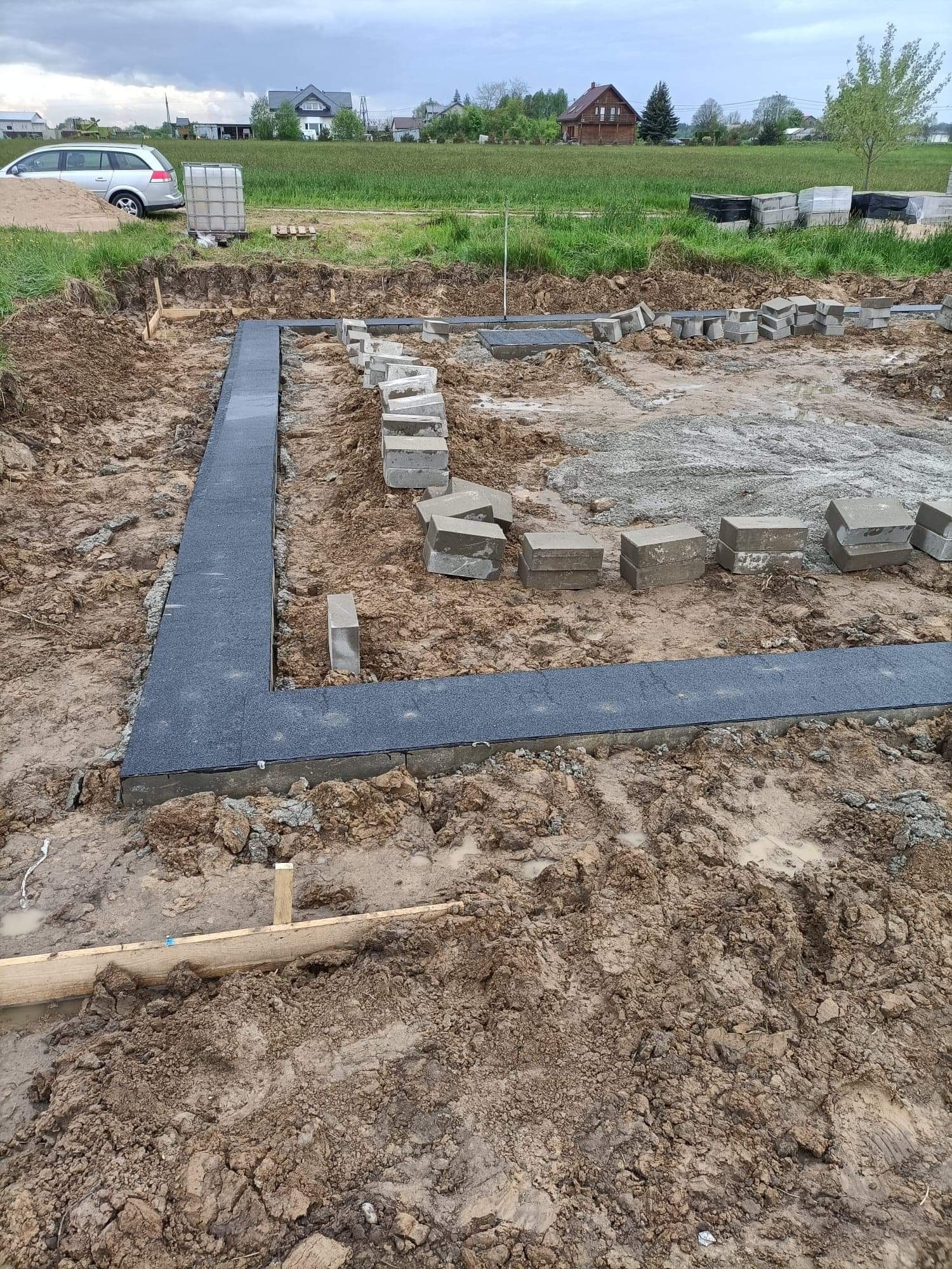 Wykop pod fundamenty domu jednorodzinnego z częściowo ułożoną izolacją poziomą w postaci szarych płyt gumowych, obok ułożone bloczki fundamentowe, w tle samochód osobowy i budynki mieszkalne.