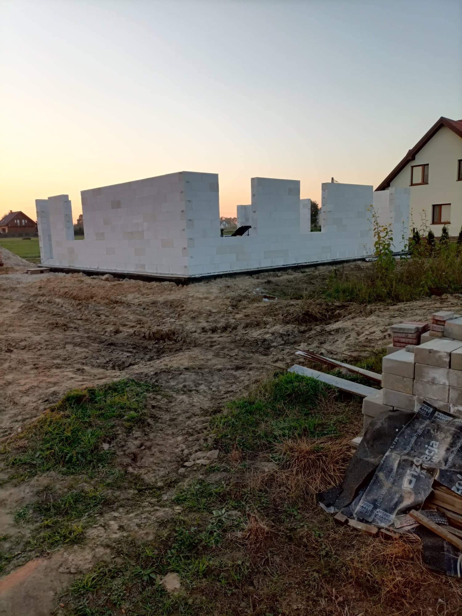 Wznoszone ściany parteru z białych bloczków betonu komórkowego na fundamencie, widoczne z ziemi, w tle dom jednorodzinny i zachodzące słońce.