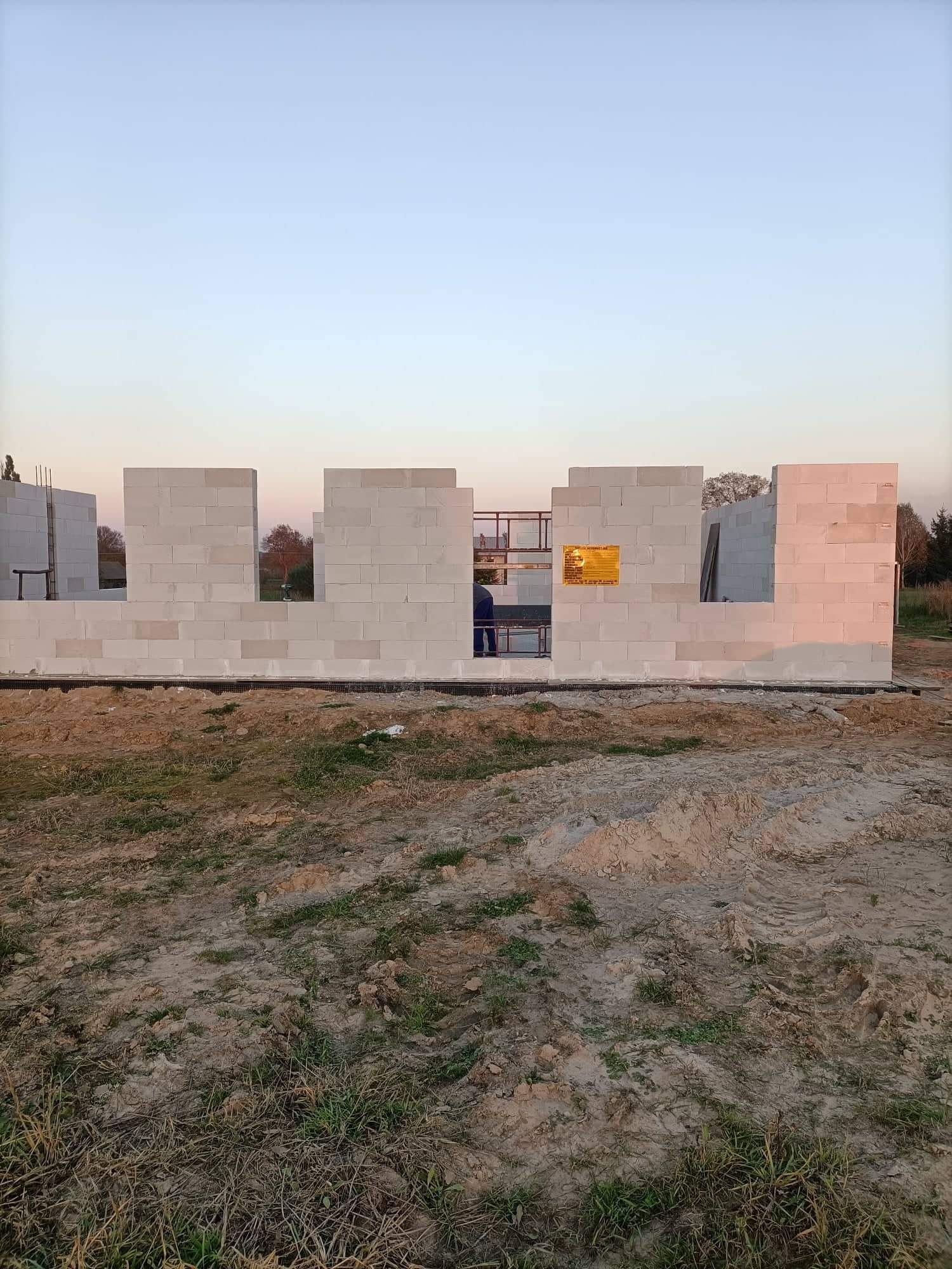 Wznoszone ściany domu z białych bloczków, widoczny pracownik wewnątrz konstrukcji i żółta tablica informacyjna na ścianie.