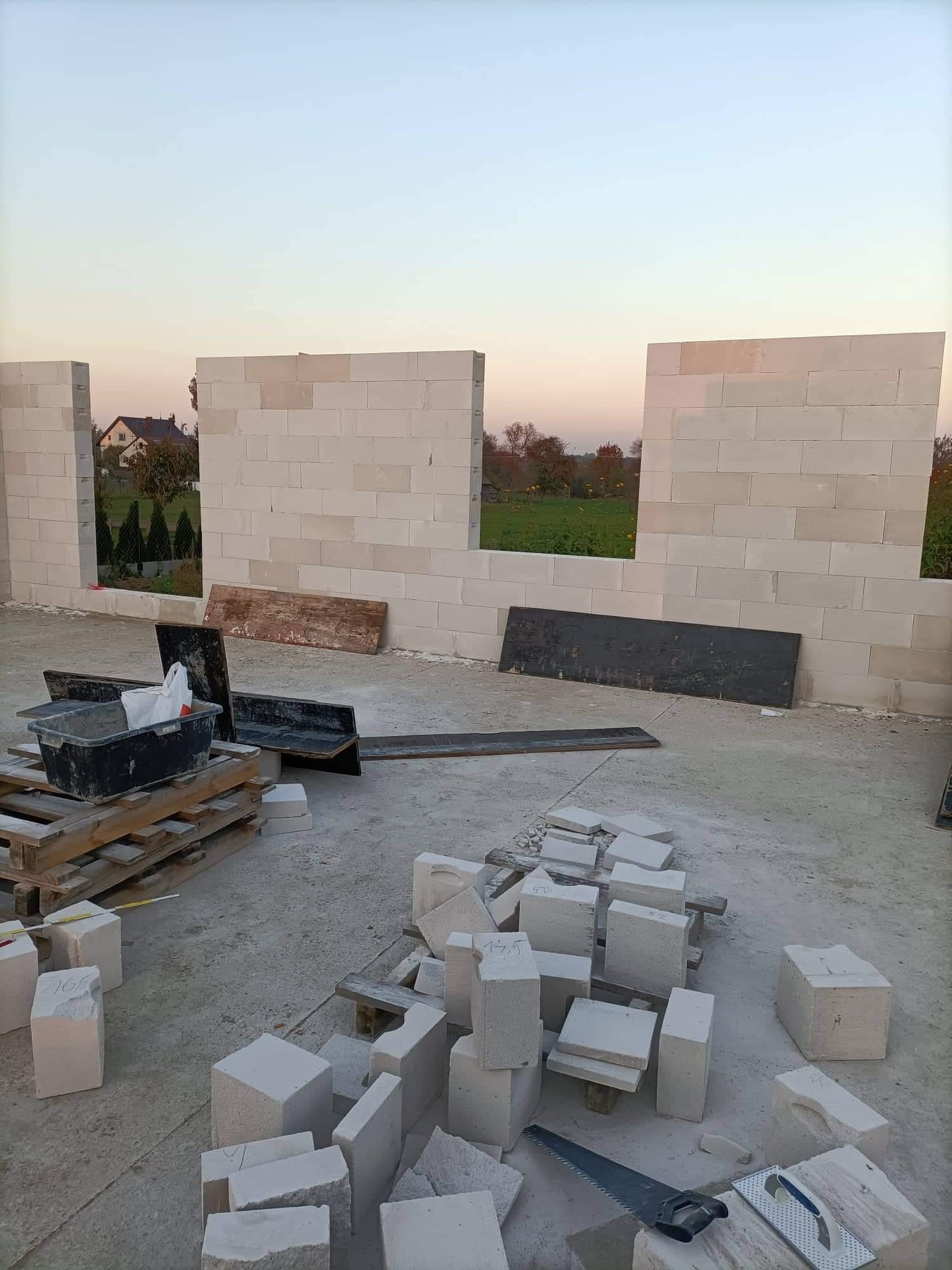 Wznoszenie ścian domu z bloczków betonu komórkowego; widoczne otwory okienne i drzwiowe; narzędzia murarskie i paleta z zaprawą w tle.