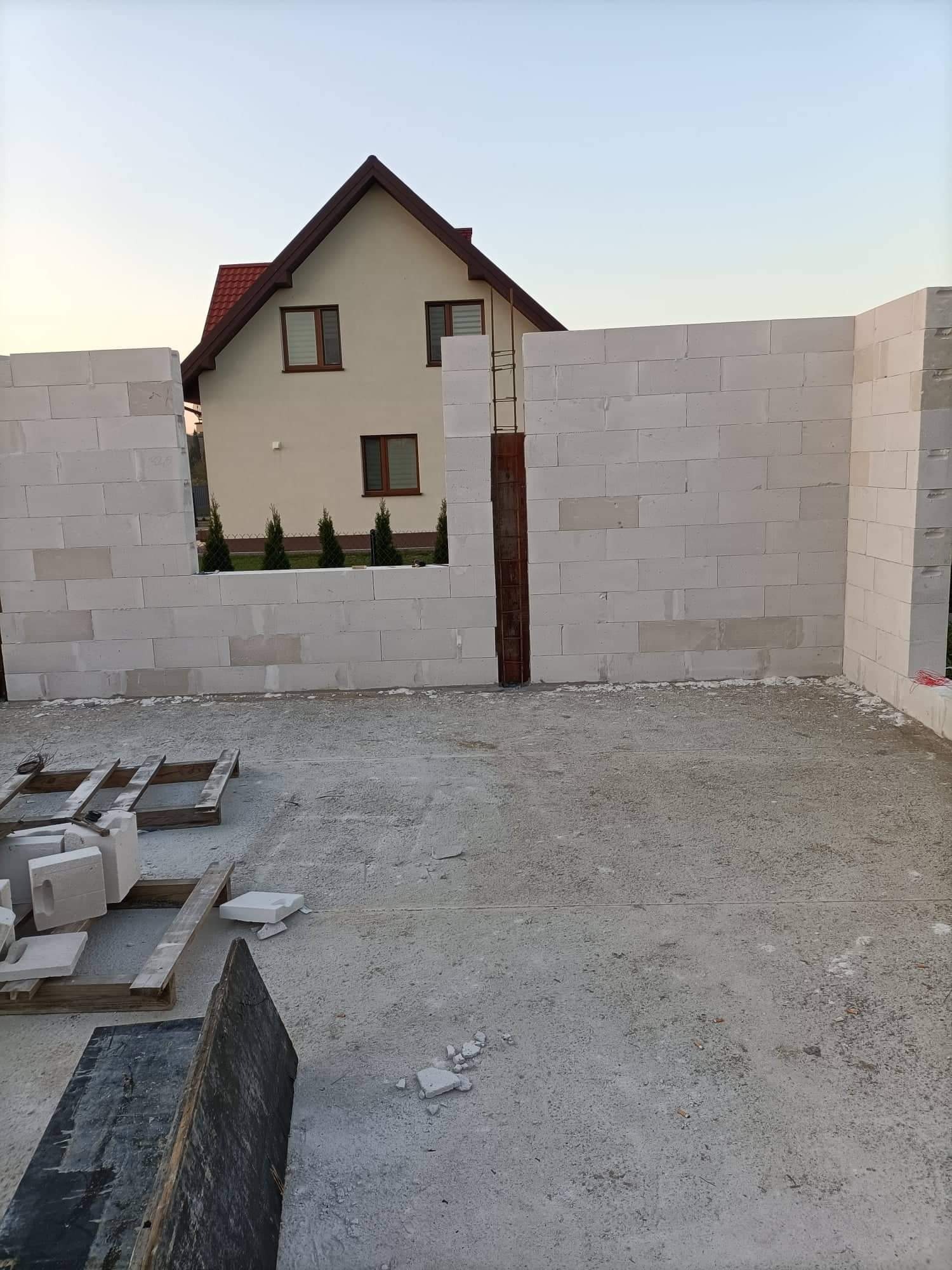 Wznoszenie ścian z białych bloczków betonu komórkowego na tle dwukondygnacyjnego domu z czerwonym dachem i zielonymi tujami w ogrodzie, widoczne drewniane palety z bloczkami i fragmenty gruzu...