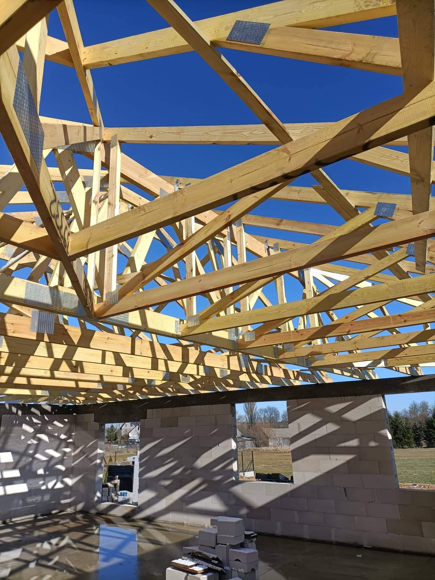 Wnętrze surowego domu z pustaków z widoczną konstrukcją drewnianą dachu na tle błękitnego nieba, elementy złączone metalowymi płytkami.