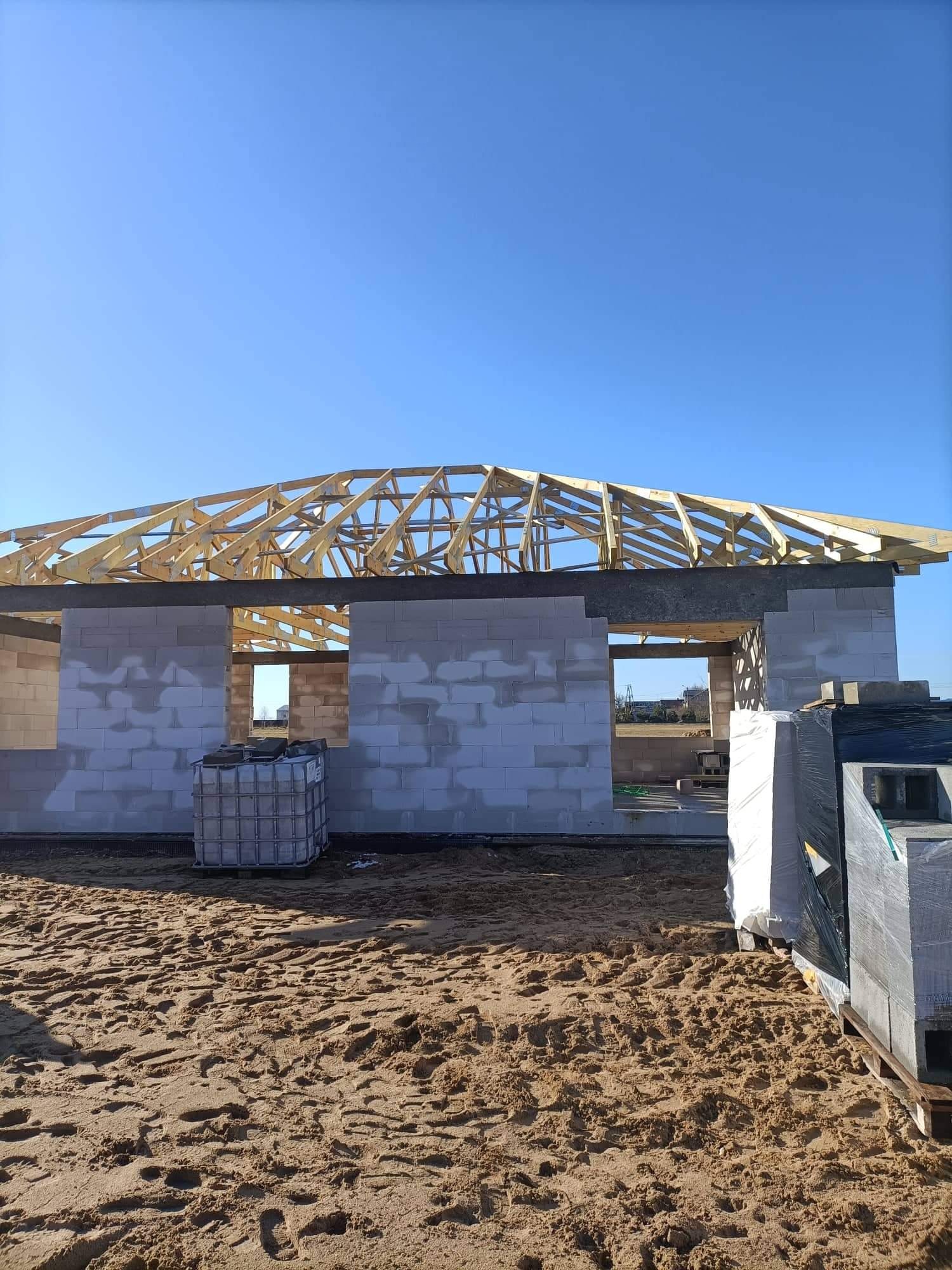 Wznoszenie domu murowanego z bloczków betonu komórkowego, widoczna drewniana konstrukcja dachu, palety z materiałem budowlanym na piaszczystym podłożu