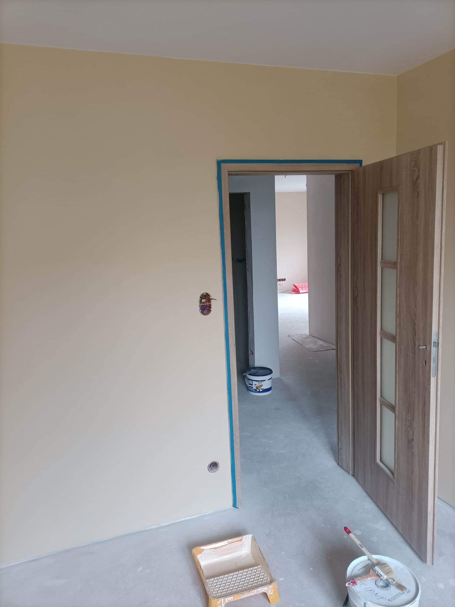 Wnętrze po malowaniu ścian na jasny kremowy kolor, widoczna otwarta drewniana ościeżnica z drzwiami, zabezpieczona taśmą malarską, puszka z farbą i wałek malarski na podłodze.