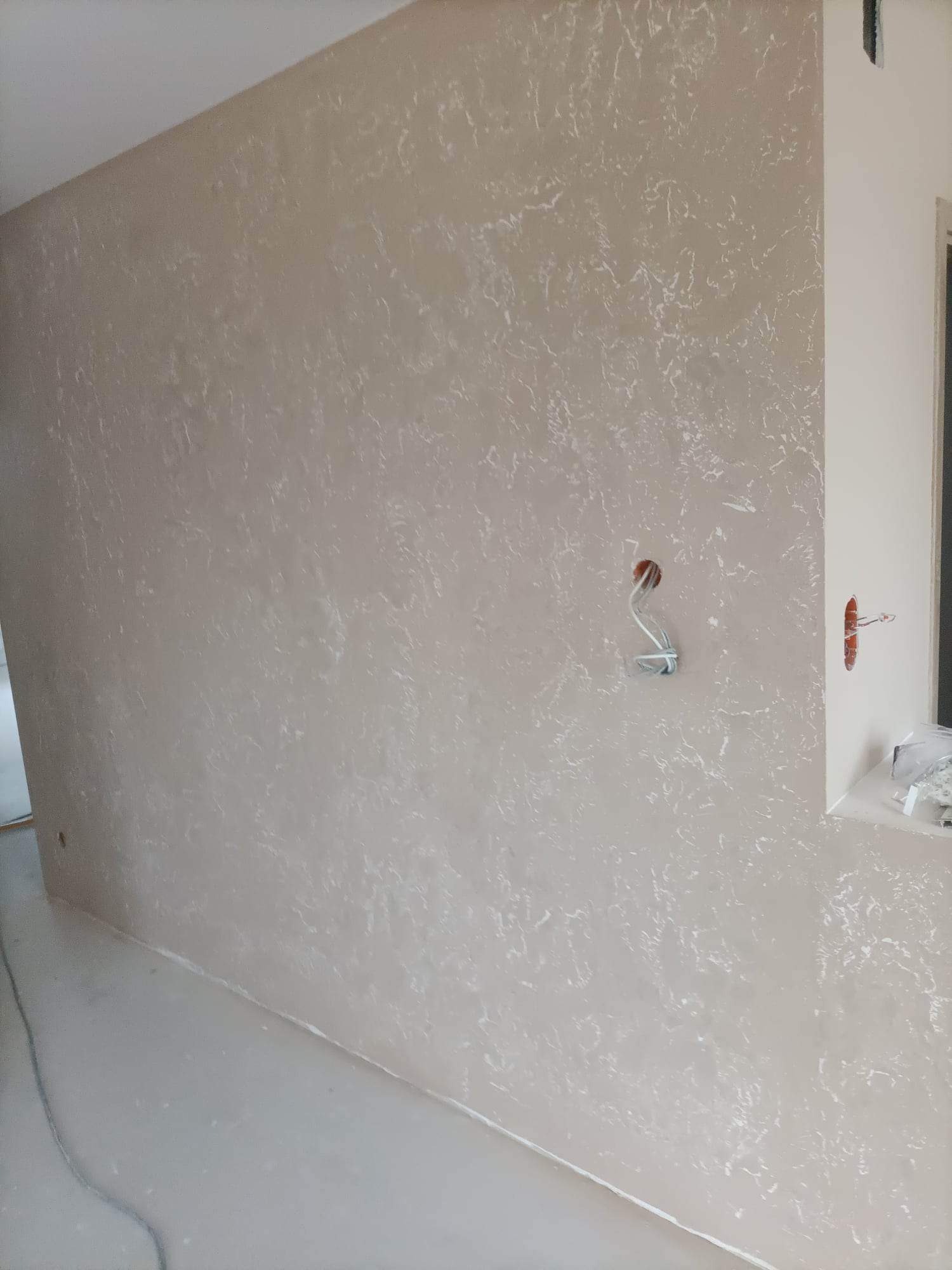 Beżowa ściana wewnętrzna z dekoracyjnym wzorem imitującym marmur, widoczne wystające przewody elektryczne i gniazdka w trakcie prac wykończeniowych.