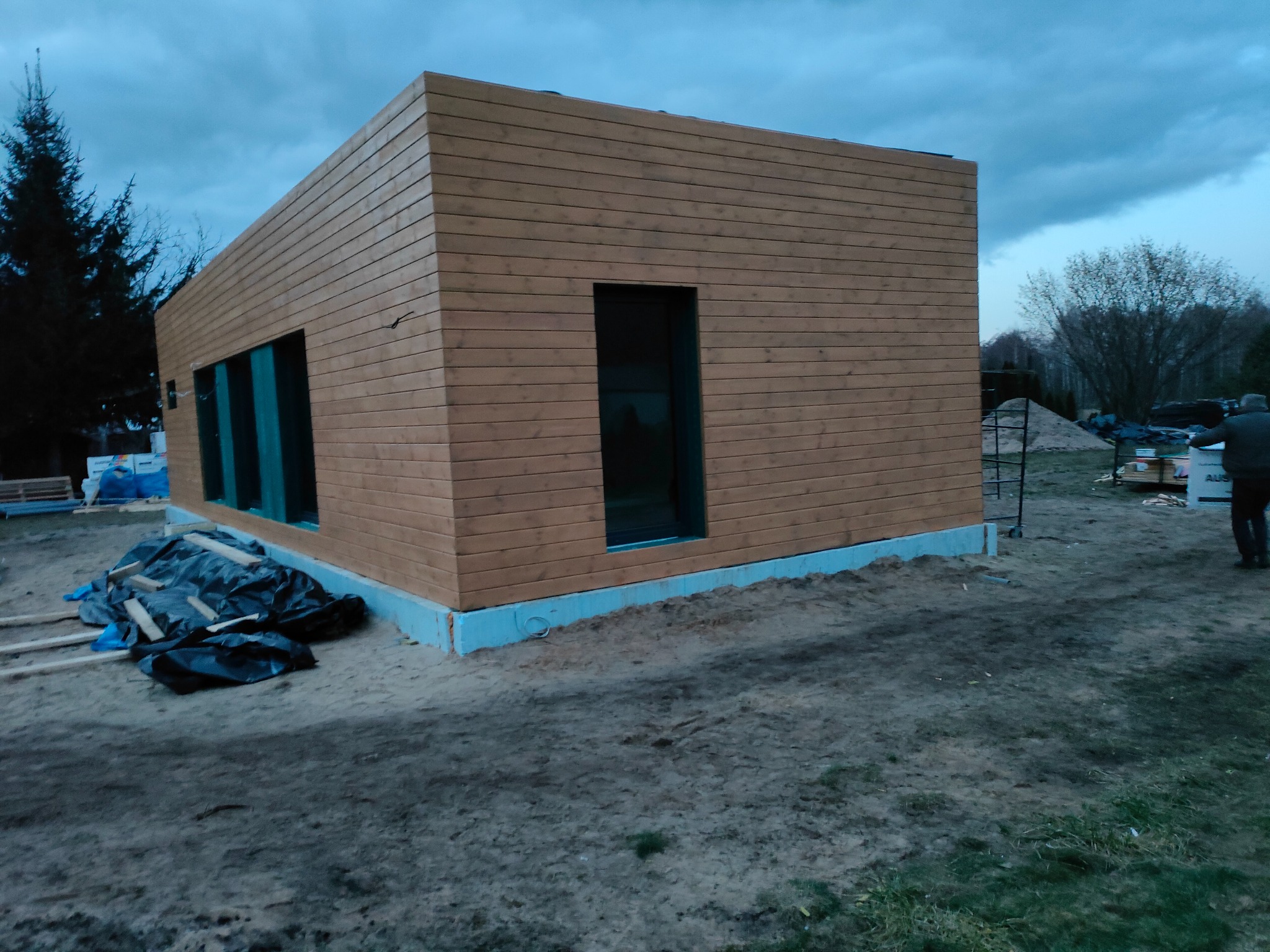 Nowoczesny, parterowy dom z drewnianą elewacją w trakcie budowy, z widocznymi oknami i fundamentami. Na pierwszym planie teren budowy z materiałami.