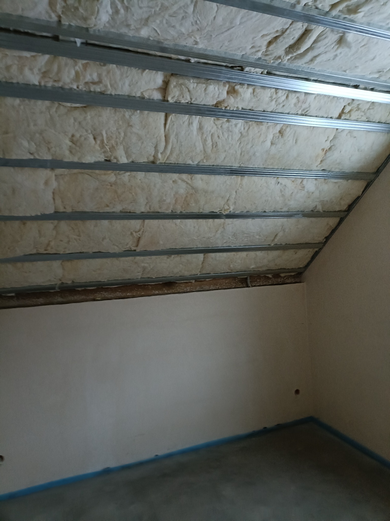 Skośne poddasze z izolacją z wełny mineralnej, widoczne metalowe profile konstrukcyjne. Częściowo otynkowana ściana i surowa podłoga. Etap prac wykończeniowych.