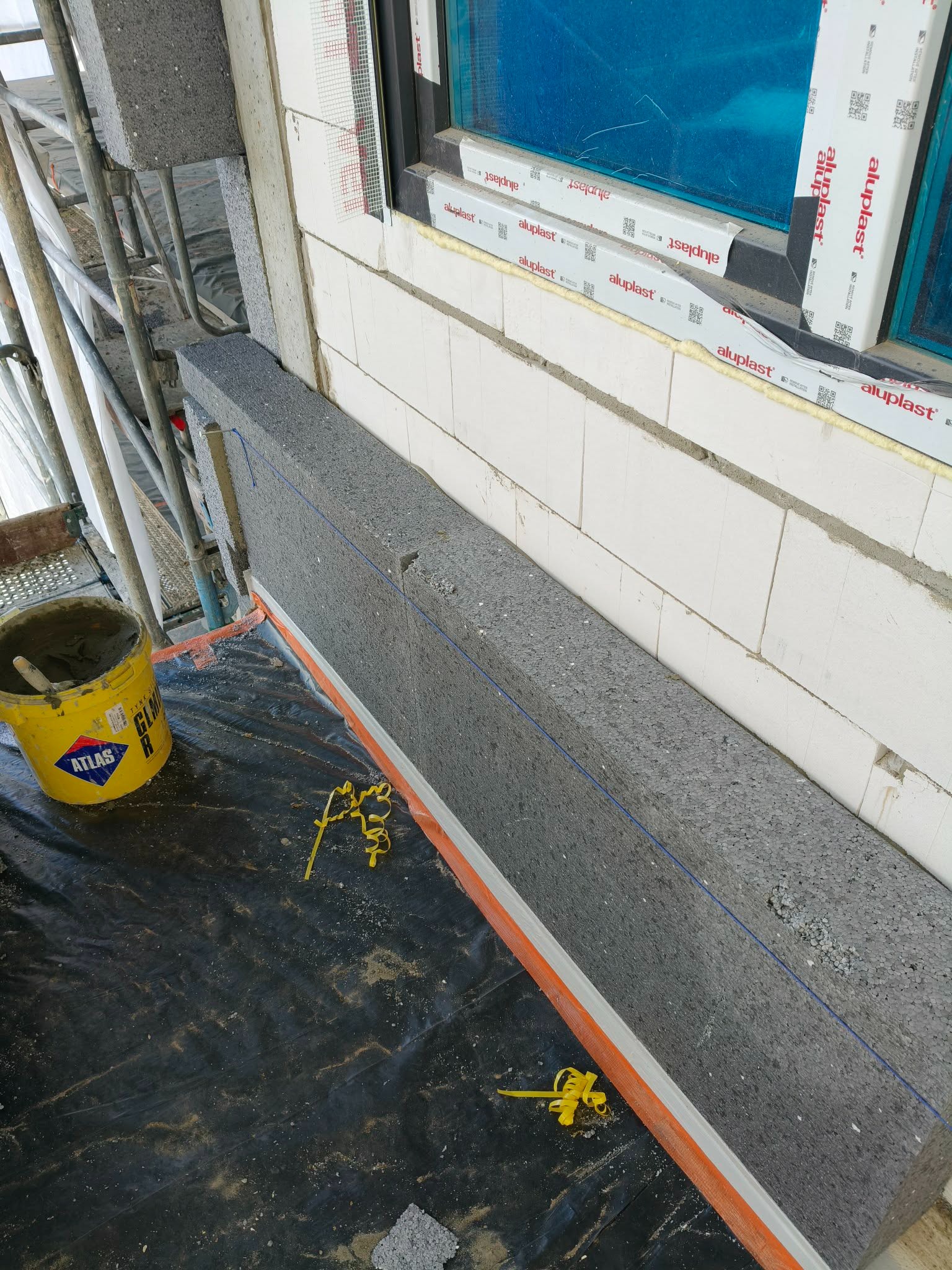 Montaż izolacji termicznej z szarego styropianu na ścianie z białych bloczków, widoczny fragment okna z folią ochronną i wiadro z zaprawą Atlas.