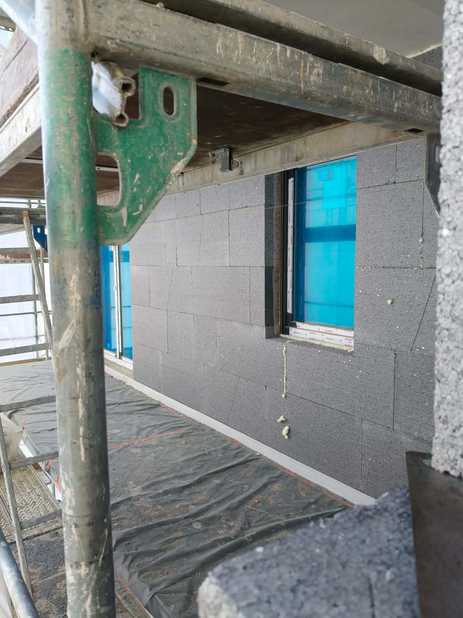 Szara ściana z bloków izolacyjnych, okno zabezpieczone folią, widok z rusztowania. Etap budowy domu z izolacją termiczną, detale montażu.